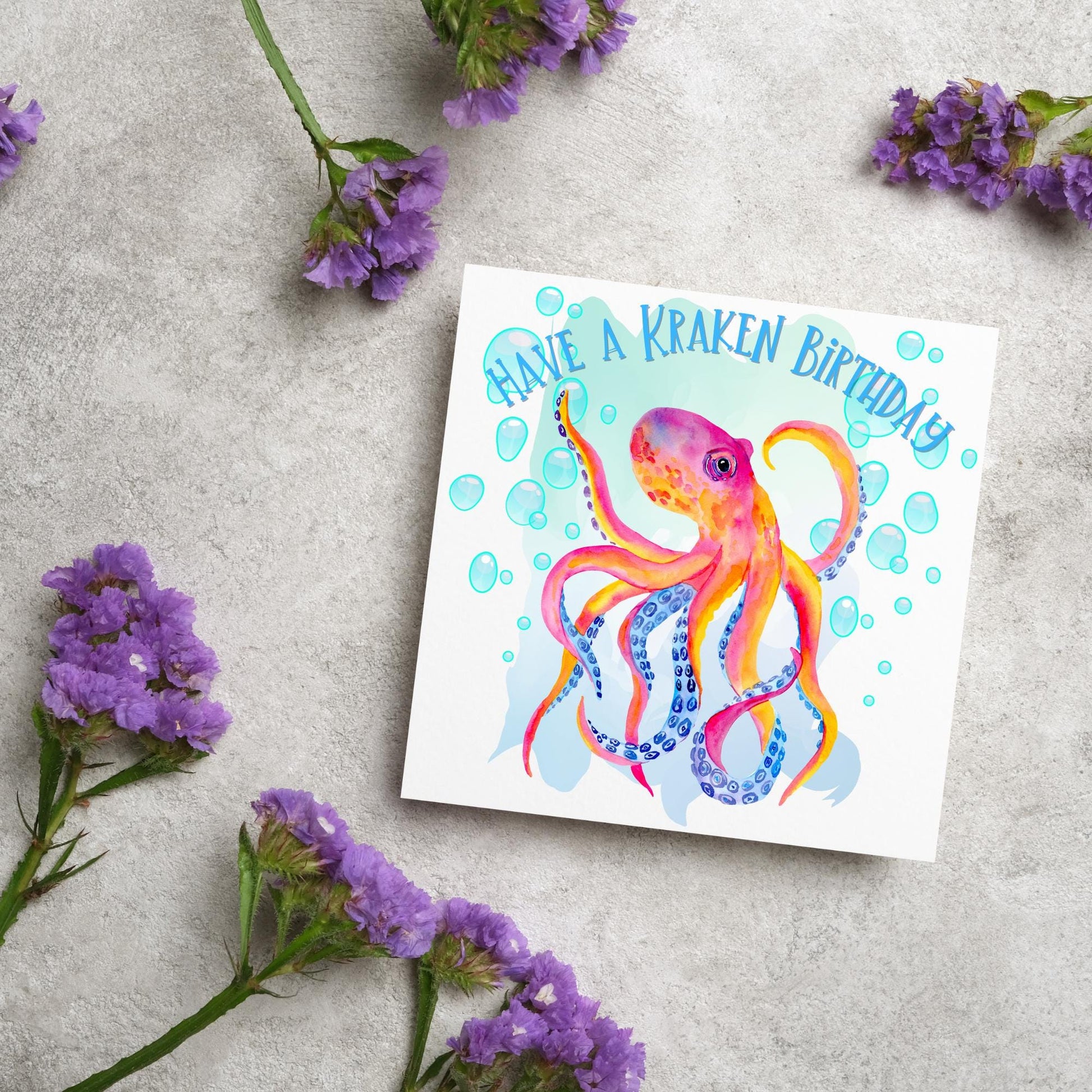 Image of a square birthday card. The background of the card is white and in the centre is a large watercolour image of an octopus in red, orange and blue tones against a light blue bubbly backdrop. Above the Octopus in blue capitalised text, have a kraken birthday is written in a handwritten style font