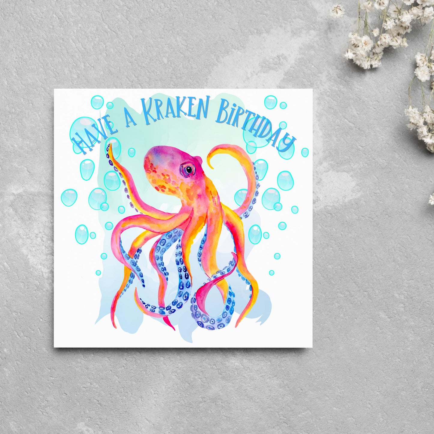 Image of a square birthday card. The background of the card is white and in the centre is a large watercolour image of an octopus in red, orange and blue tones against a light blue bubbly backdrop. Above the Octopus in blue capitalised text, have a kraken birthday is written in a handwritten style font