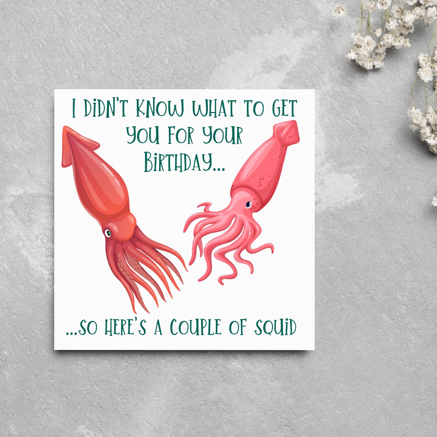 Image of a square birthday card. The background of the card is white and there is an illustration of 2 red squid in the centre. Writing surrounds the picture with 'I didn't know what to get you for your birthday....so here's a couple of squid' in a green capitalised handwritten font.