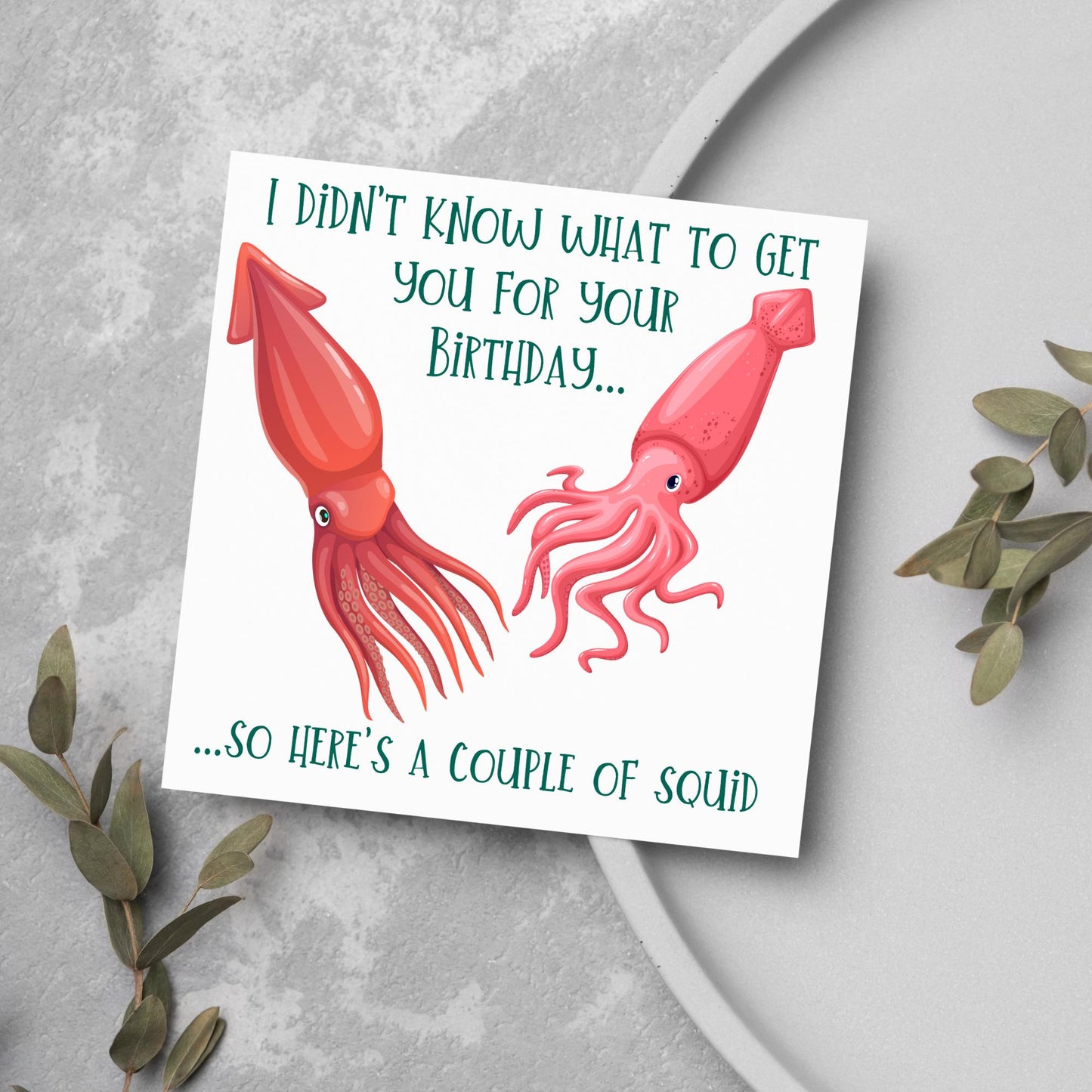 Image of a square birthday card. The background of the card is white and there is an illustration of 2 red squid in the centre. Writing surrounds the picture with 'I didn't know what to get you for your birthday....so here's a couple of squid' in a green capitalised handwritten font.