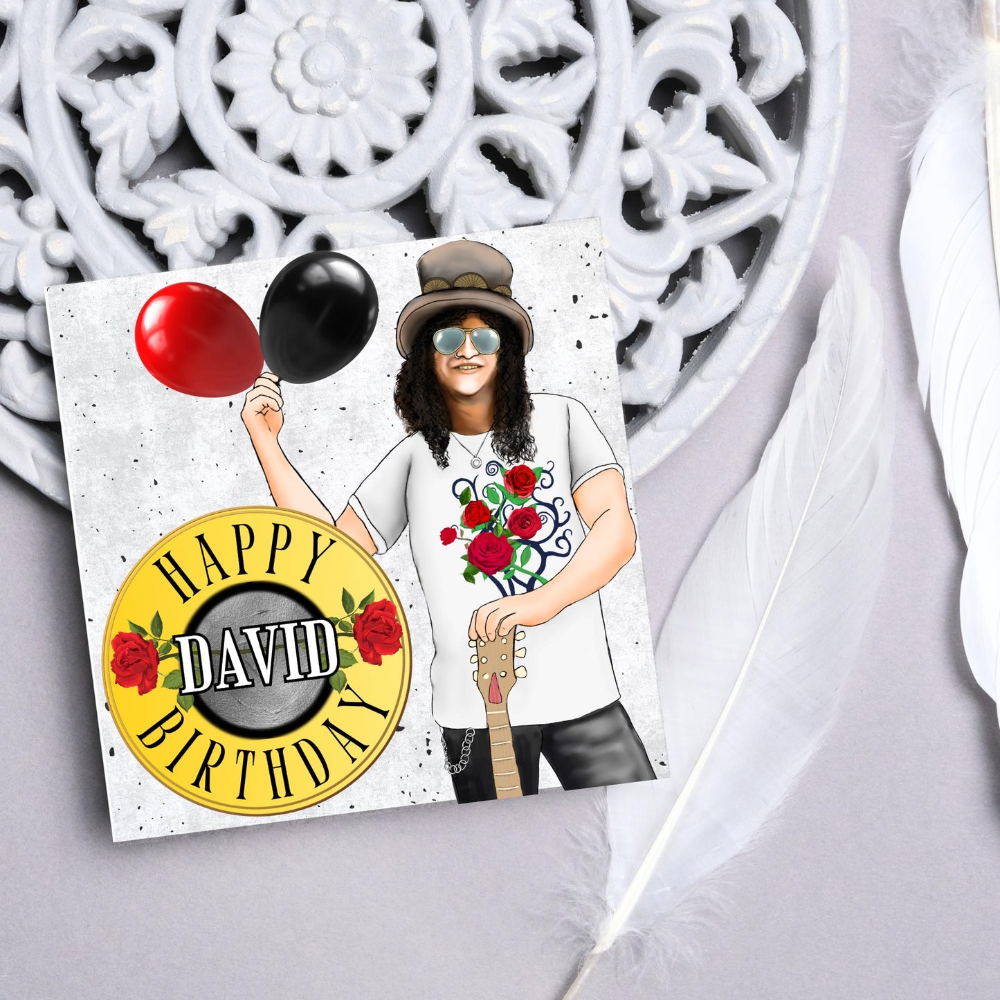 Square Birthday card with an illustration of Slash from Guns and Roses holding a red balloon and a black balloon in one hand, and resting his other on his guitar. He is wearing his trademark top hat, a rose print tee shirt and sunglasses. There is a band style logo that reads happy birthday david to the left of him.