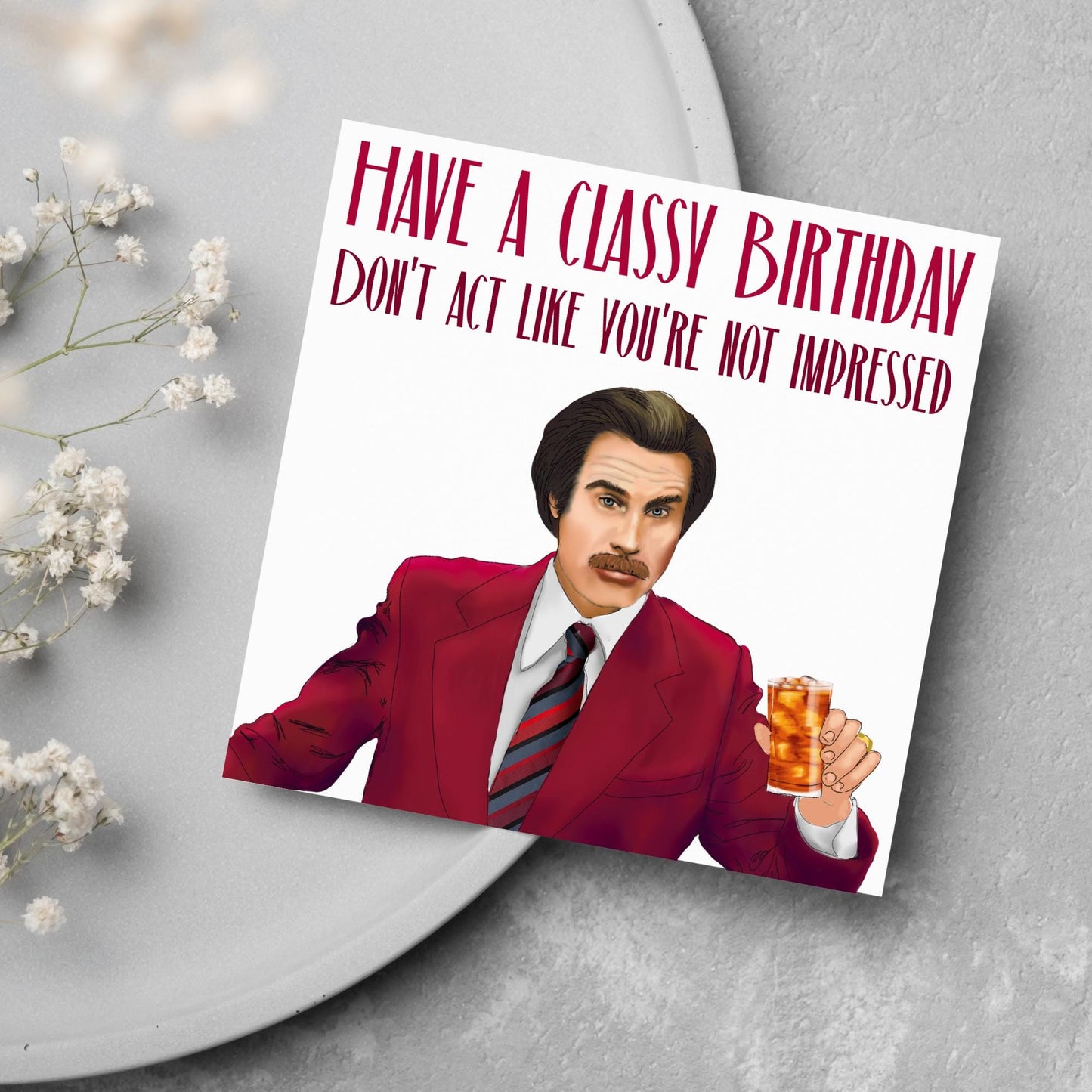 image of a square birthday card, the card has a white background with a hand drawn illustration of Ron Burgandy from the Movie Anchorman in a dark red suit holding a glass of whiskey. Above him is dark red capitalised text that reads 'Have a classy birthday, Don't act like you're not impressed.'