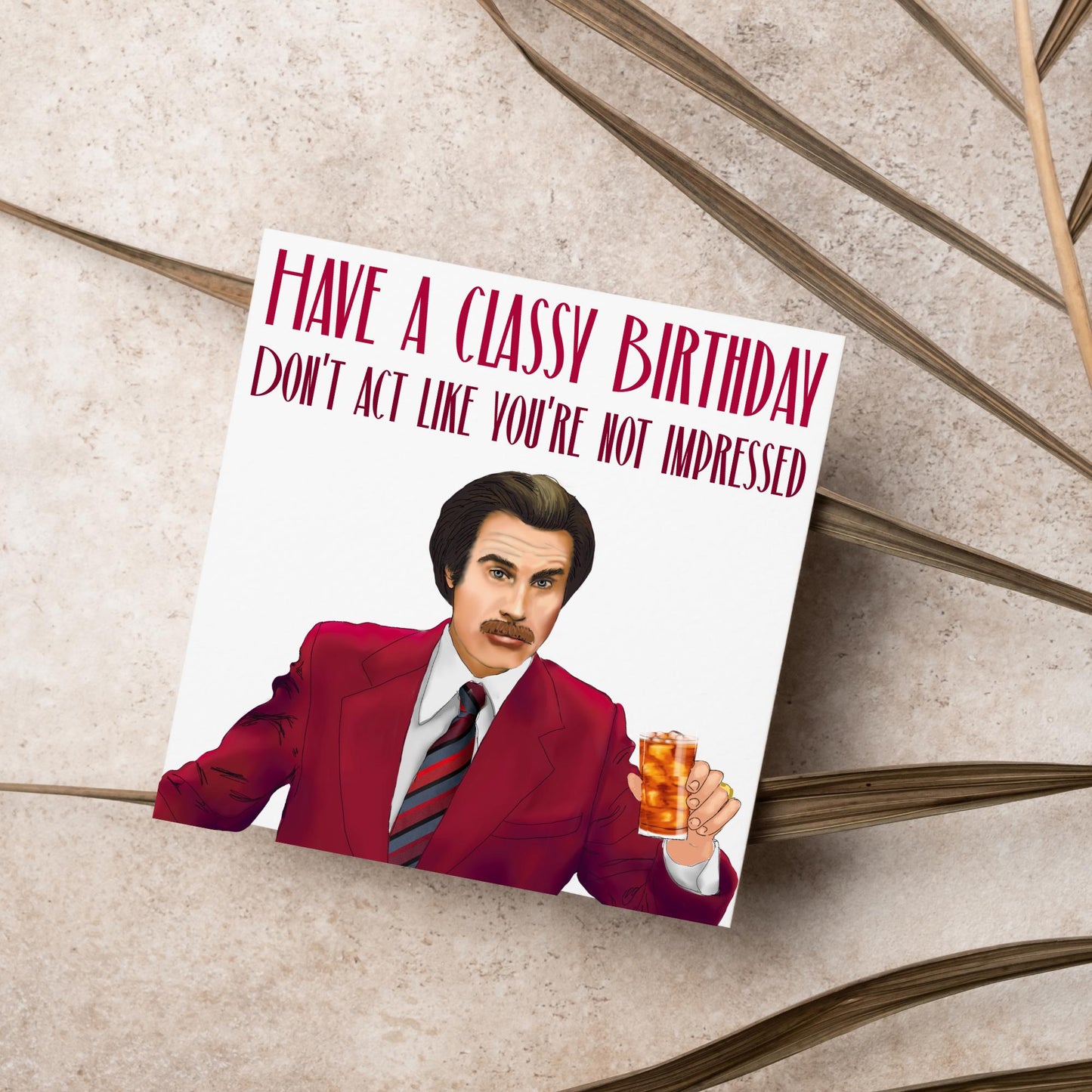 image of a square birthday card, the card has a white background with a hand drawn illustration of Ron Burgandy from the Movie Anchorman in a dark red suit holding a glass of whiskey. Above him is dark red capitalised text that reads 'Have a classy birthday, Don't act like you're not impressed.'