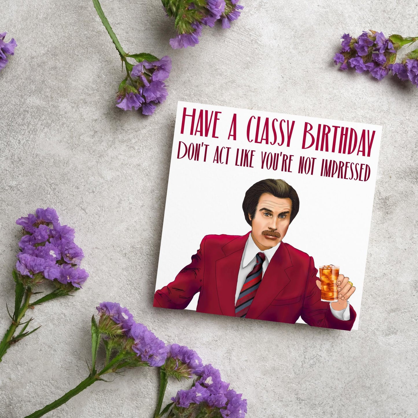 image of a square birthday card, the card has a white background with a hand drawn illustration of Ron Burgandy from the Movie Anchorman in a dark red suit holding a glass of whiskey. Above him is dark red capitalised text that reads 'Have a classy birthday, Don't act like you're not impressed.'