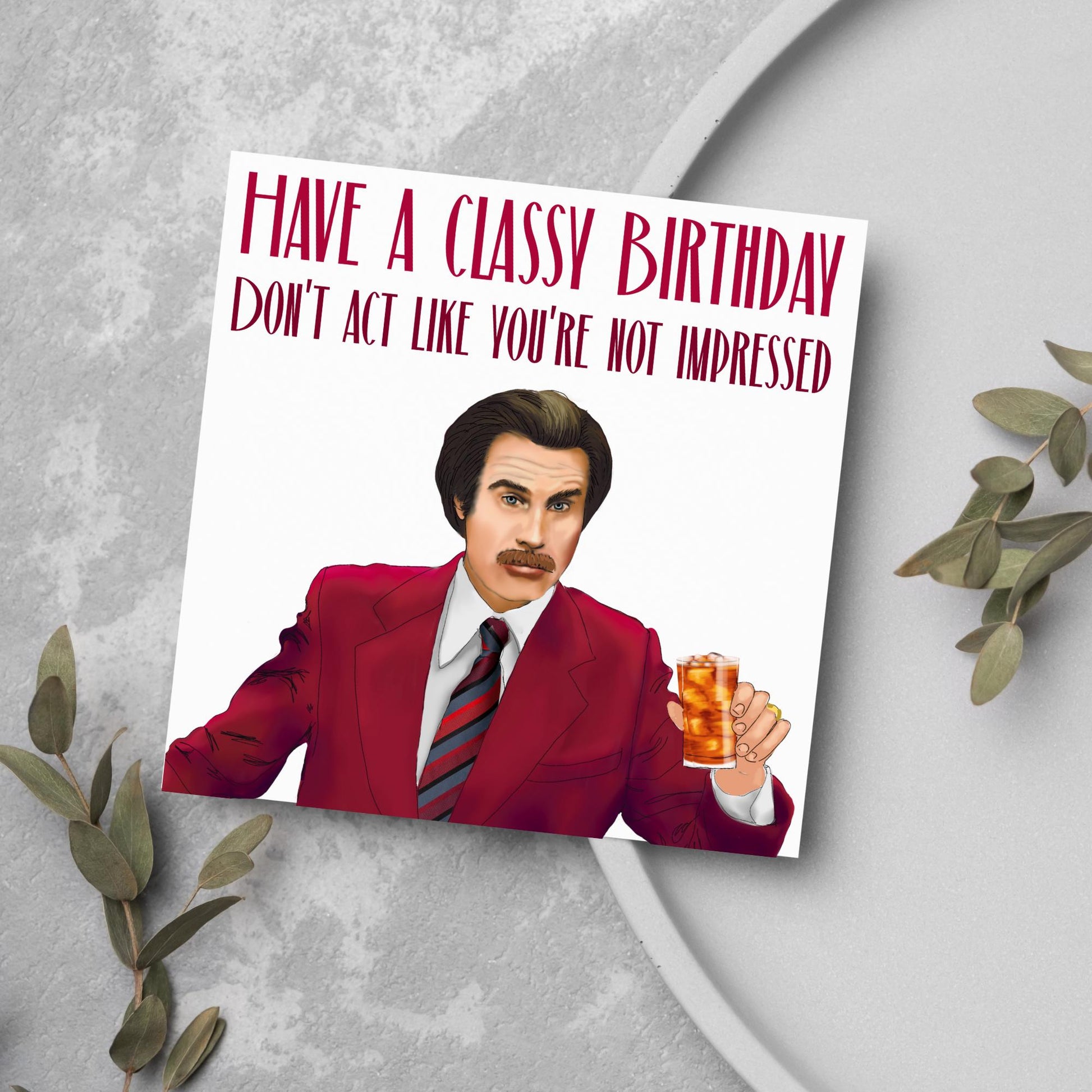 image of a square birthday card, the card has a white background with a hand drawn illustration of Ron Burgandy from the Movie Anchorman  in a dark red suit holding a glass of whiskey. Above him is dark red capitalised text that reads 'Have a classy birthday, Don't act like you're not impressed.'