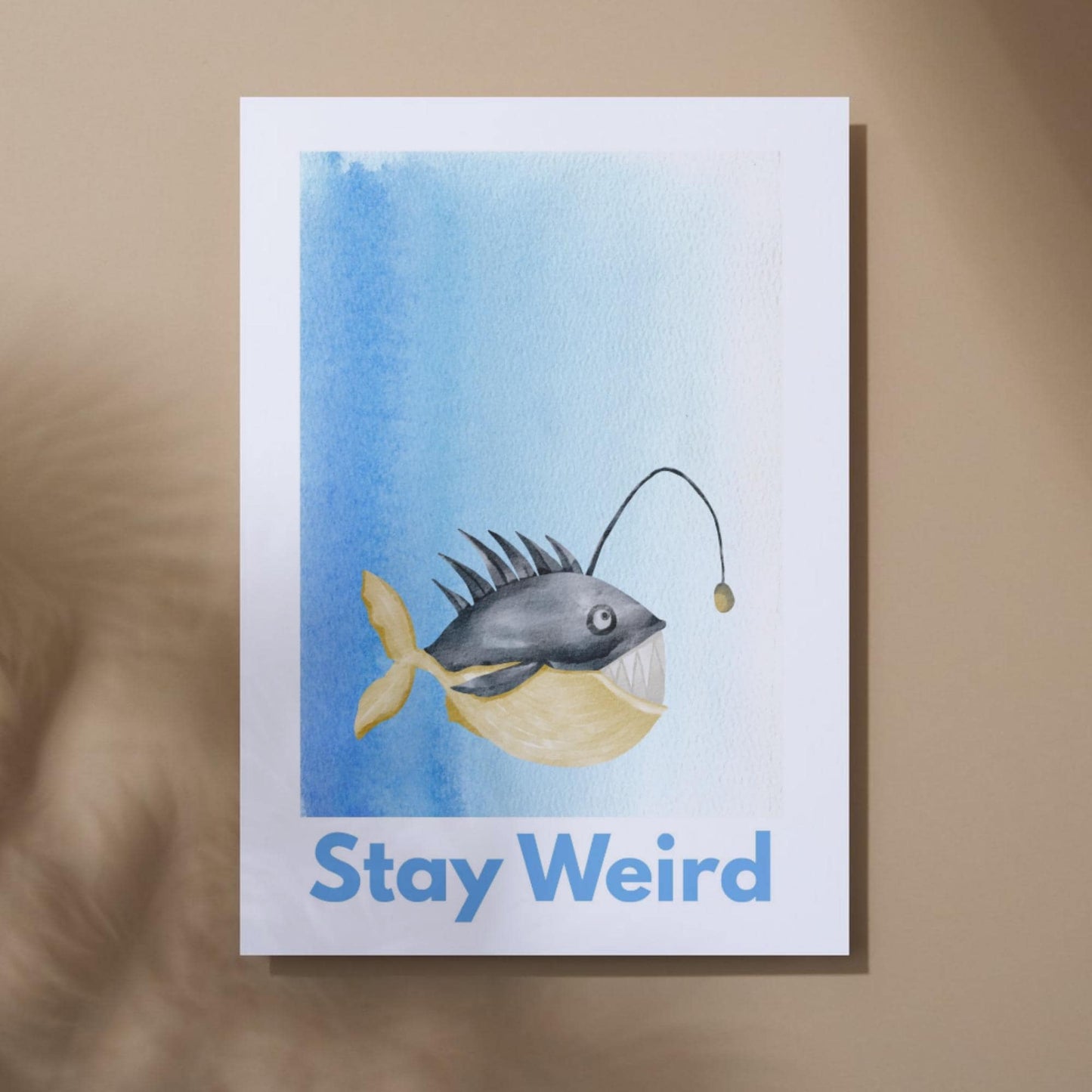 An A4 art print featuring a whimsical watercolor illustration of an anglerfish with a yellow tail and light lure, set against a blue gradient background. Below the fish, bold blue text reads "Stay Weird."