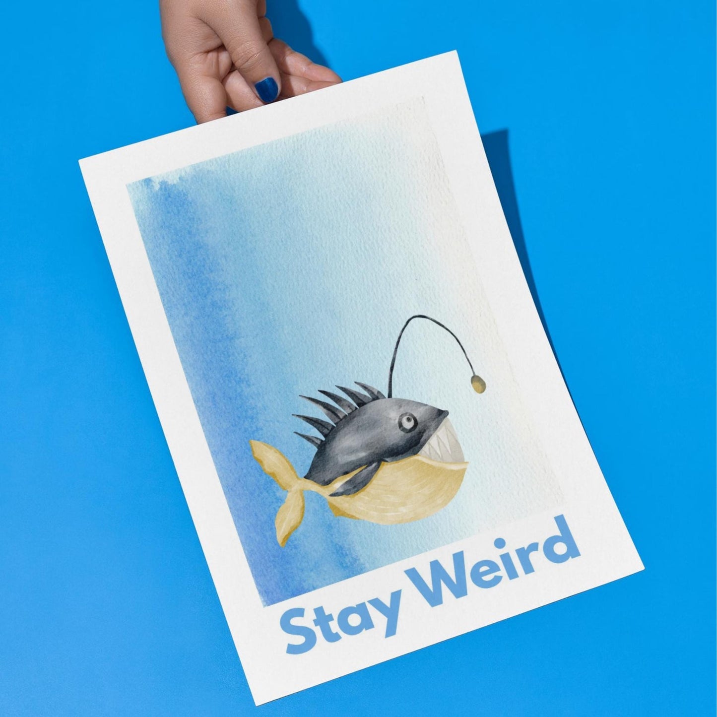 An A4 art print featuring a whimsical watercolor illustration of an anglerfish with a yellow tail and light lure, set against a blue gradient background. Below the fish, bold blue text reads "Stay Weird."