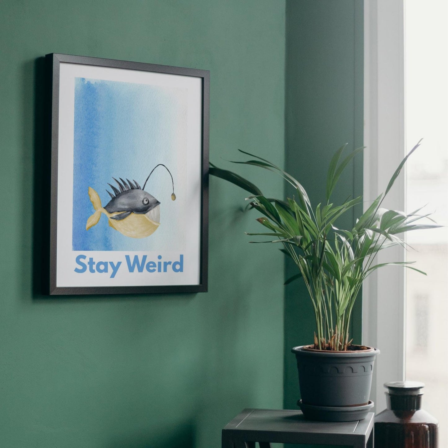 A framed A4 art print featuring a whimsical watercolor illustration of an anglerfish with a yellow tail and light lure, set against a blue gradient background. Below the fish, bold blue text reads "Stay Weird."