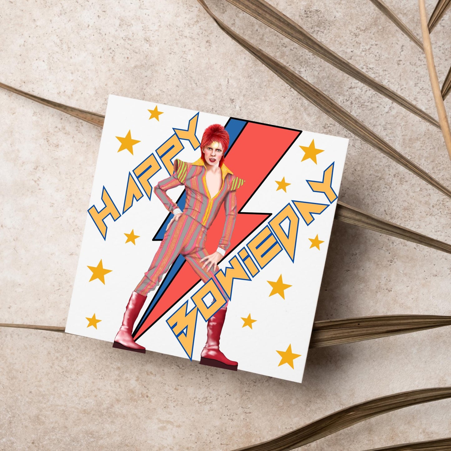 image of a square birthday card. Card has a white background with a vibrant illustration of David bowie with red hair and wearing a striped jumpsuit, behind him is the iconic Bowie lightning bolt in bright red and blue. Diagonal text reads Happy Bowieday in zigzag design bold yellow font. There are yellow stars dotted around the white background.