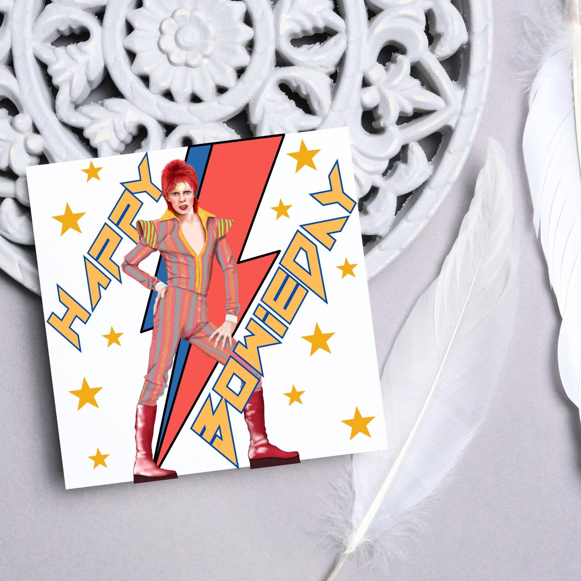 image of a square birthday card. Card has a white background with a vibrant illustration of David bowie with red hair and wearing a striped jumpsuit, behind him is the iconic Bowie lightning bolt in bright red and blue. Diagonal text reads Happy Bowieday in zigzag design bold yellow font. There are yellow stars dotted around the white background.