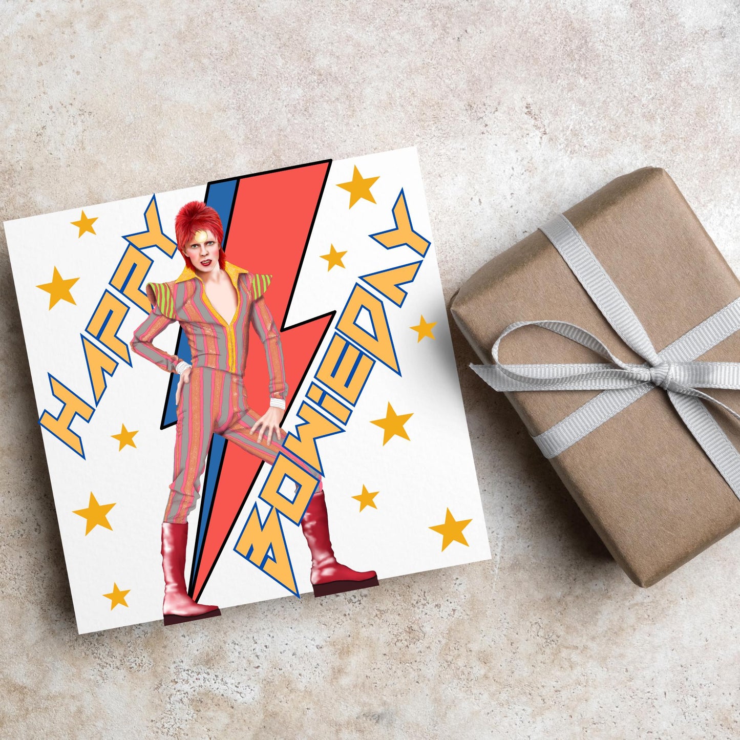 image of a square birthday card. Card has a white background with a vibrant illustration of David bowie with red hair and wearing a striped jumpsuit, behind him is the iconic Bowie lightning bolt in bright red and blue. Diagonal text reads Happy Bowieday in zigzag design bold yellow font. There are yellow stars dotted around the white background.