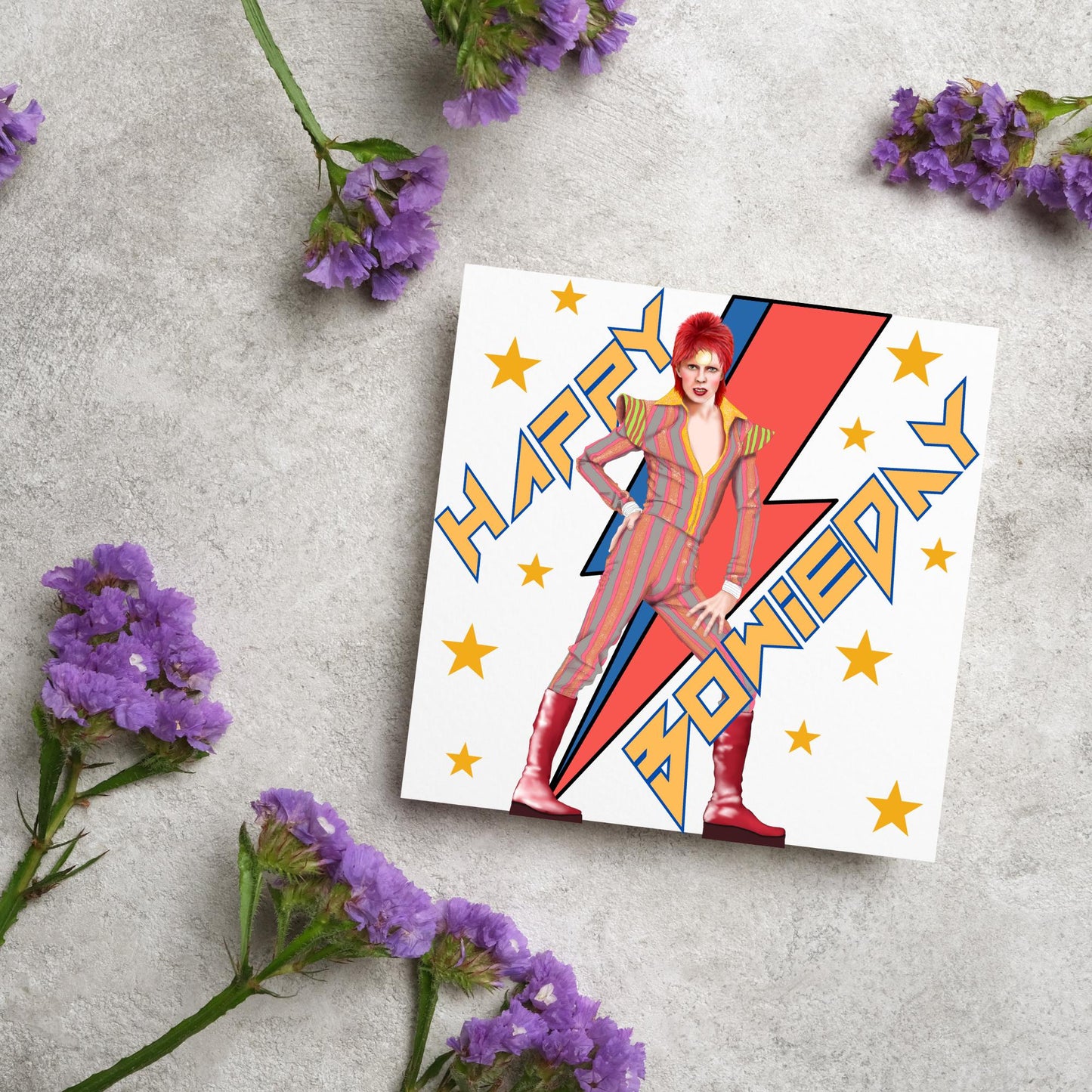 image of a square birthday card. Card has a white background with a vibrant illustration of David bowie with red hair and wearing a striped jumpsuit, behind him is the iconic Bowie lightning bolt in bright red and blue. Diagonal text reads Happy Bowieday in zigzag design bold yellow font. There are yellow stars dotted around the white background.