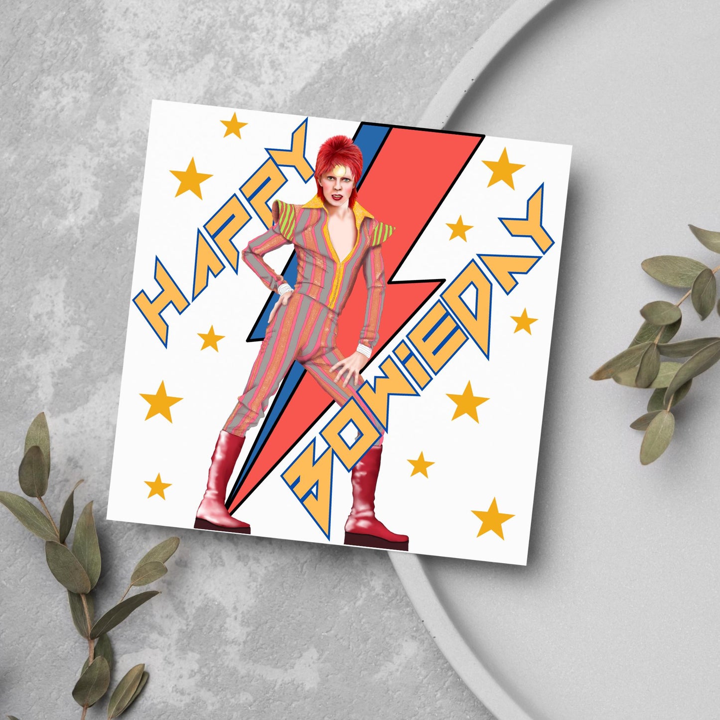 image of a square birthday card. Card has a white background with a vibrant illustration of David bowie with red hair and wearing a striped jumpsuit, behind him is the iconic Bowie lightning bolt in bright red and blue. Diagonal text reads Happy Bowieday in zigzag design bold yellow font. There are yellow stars dotted around the white background.