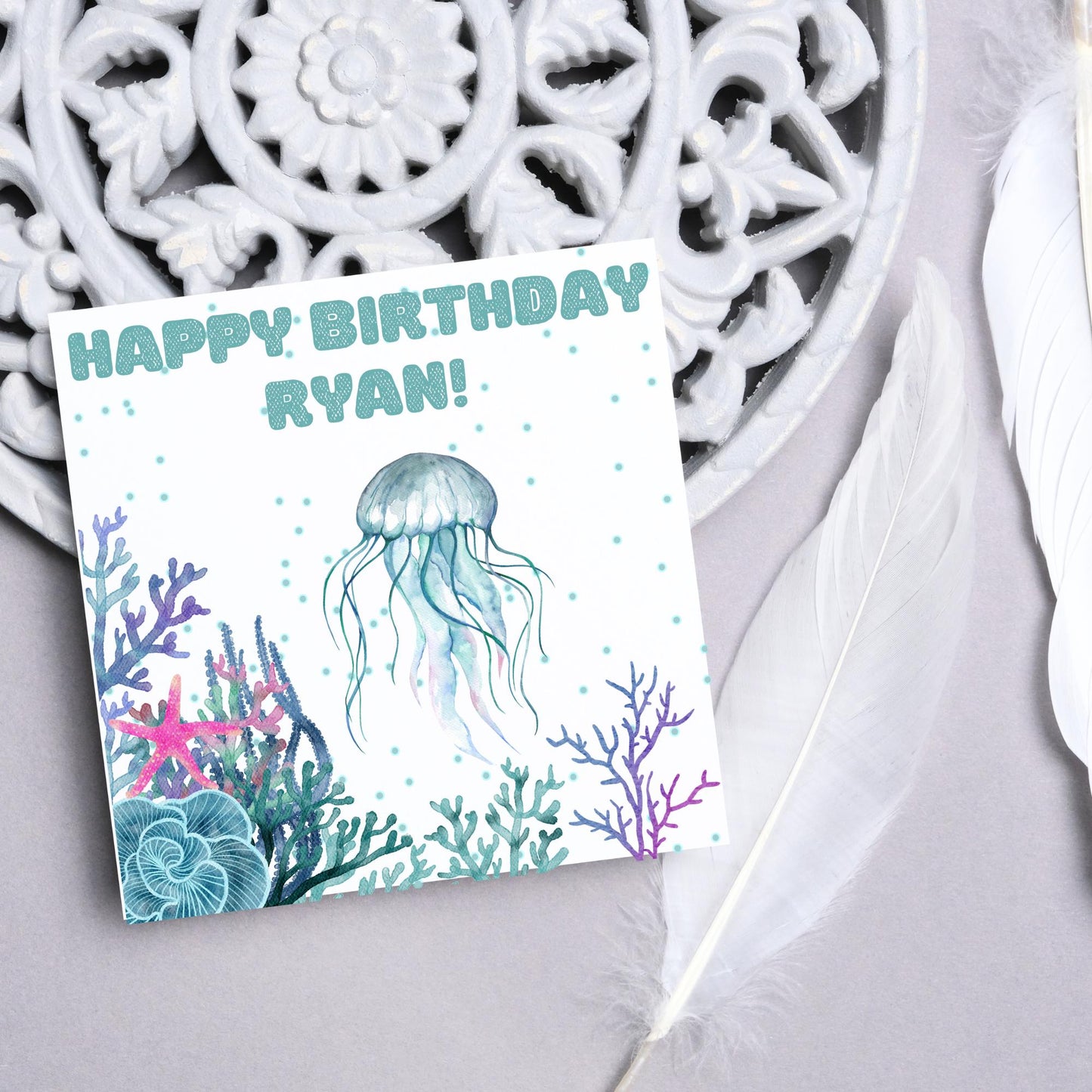 Image of a square Birthday card. Card has a white background and a watercolour style jellyfish amongst coral and seaweed. All the colours are blues, greens, pinks and purples. At the top of the card is bold blue text that says happy birthday ryan which can be personalised to any name.