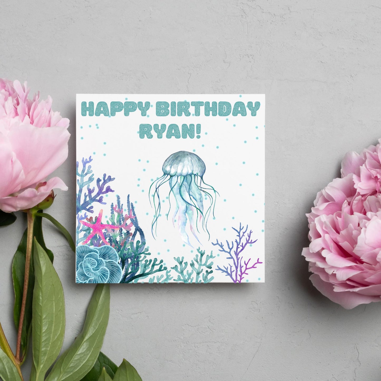 Image of a square Birthday card. Card has a white background and a watercolour style jellyfish amongst coral and seaweed. All the colours are blues, greens, pinks and purples. At the top of the card is bold blue text that says happy birthday ryan which can be personalised to any name.