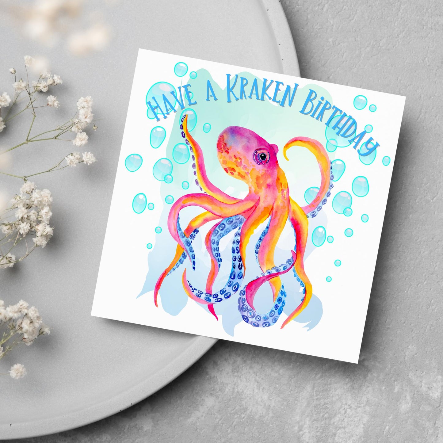Image of a square birthday card. The background of the card is white and in the centre is a large watercolour image of an octopus in red, orange and blue tones against a light blue bubbly backdrop. Above the Octopus in blue capitalised text, have a kraken birthday is written in a handwritten style font