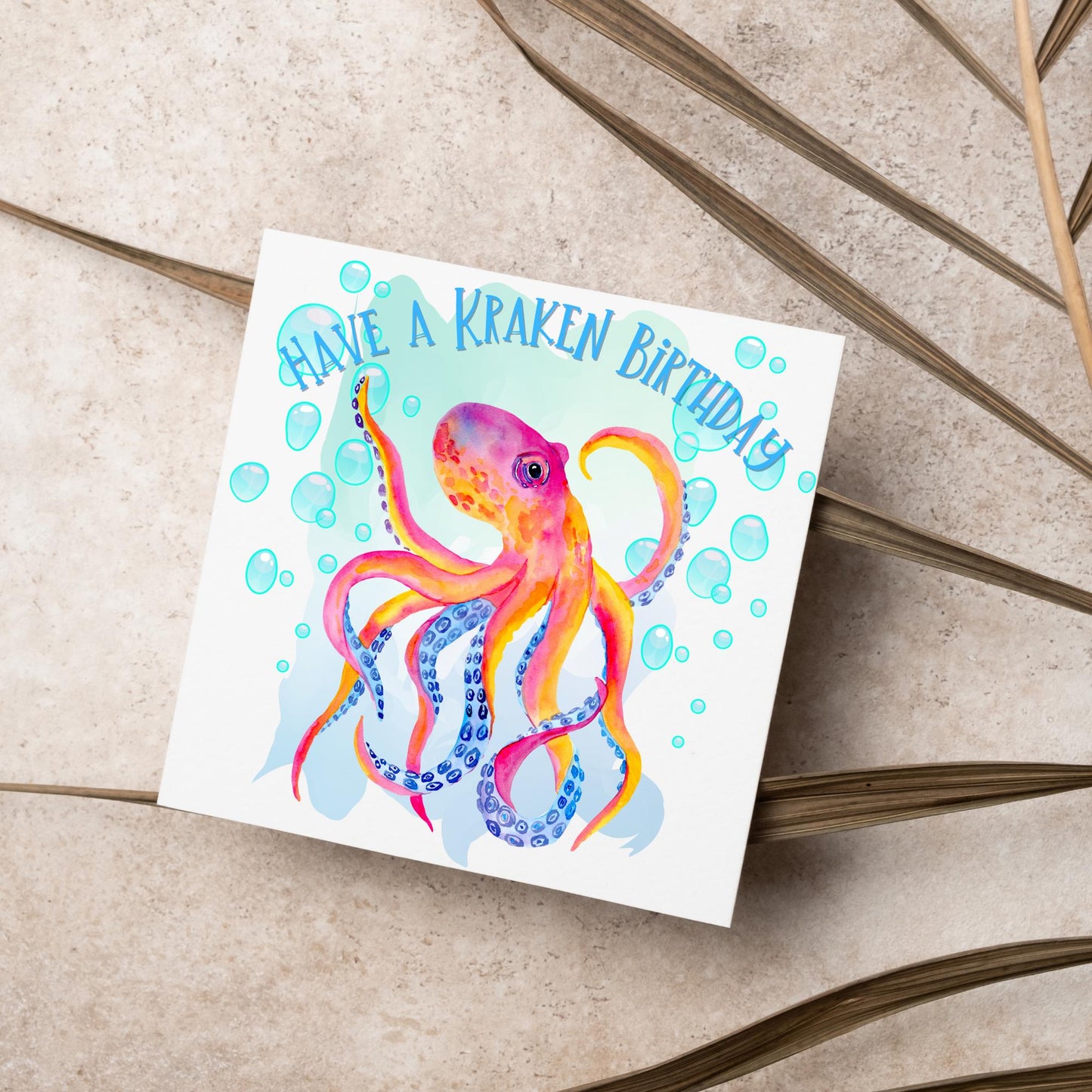 Image of a square birthday card. The background of the card is white and in the centre is a large watercolour image of an octopus in red, orange and blue tones against a light blue bubbly backdrop. Above the Octopus in blue capitalised text, have a kraken birthday is written in a handwritten style font