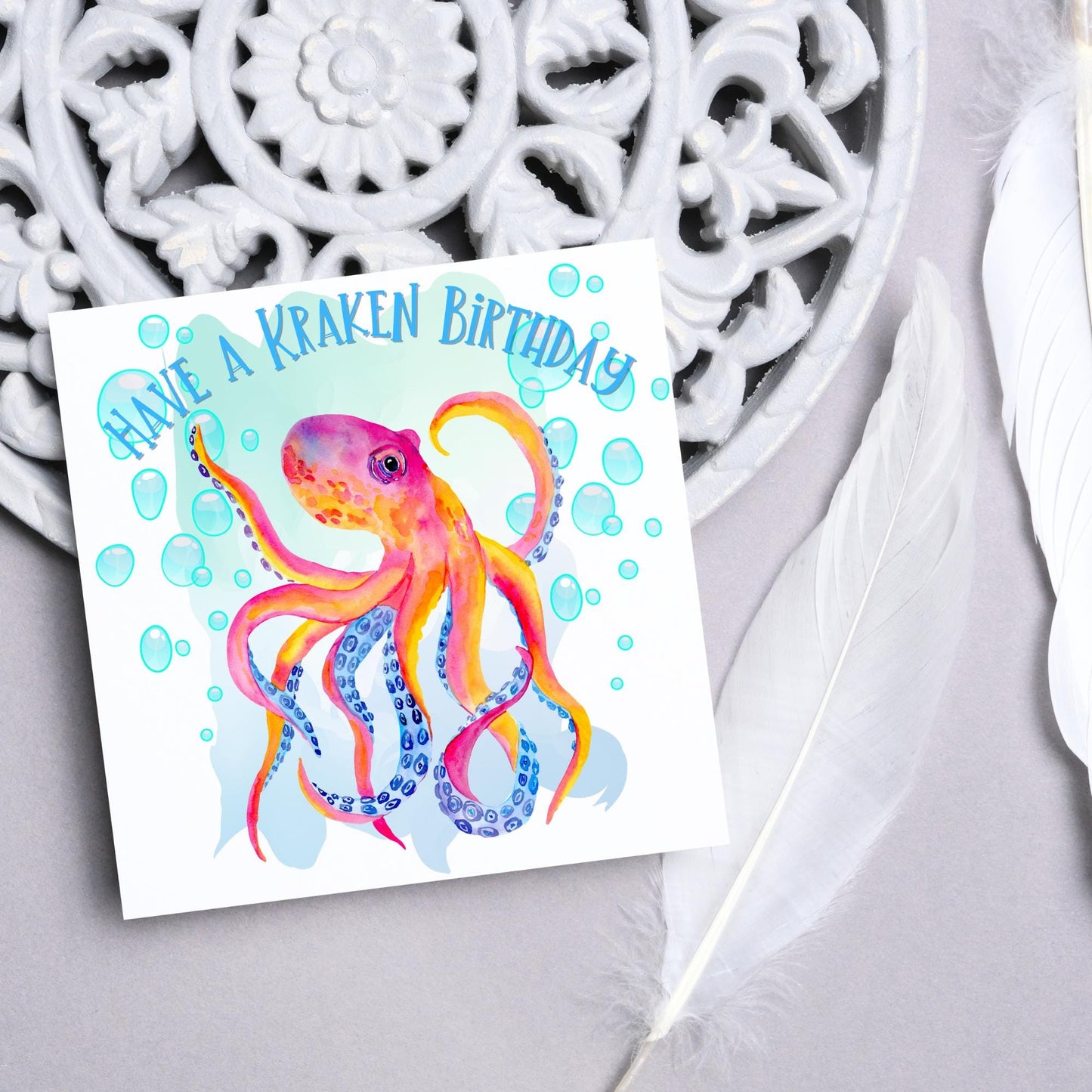 Image of a square birthday card. The background of the card is white and in the centre is a large watercolour image of an octopus in red, orange and blue tones against a light blue bubbly backdrop. Above the Octopus in blue capitalised text, have a kraken birthday is written in a handwritten style font