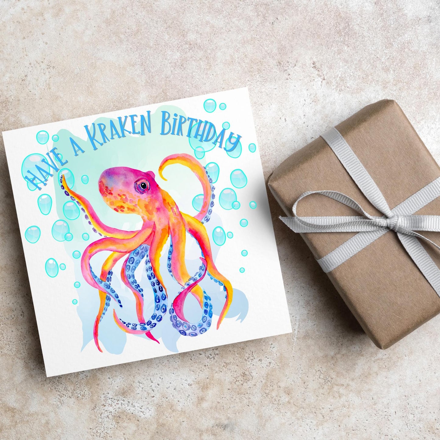 Image of a square birthday card. The background of the card is white and in the centre is a large watercolour image of an octopus in red, orange and blue tones against a light blue bubbly backdrop. Above the Octopus in blue capitalised text, have a kraken birthday is written in a handwritten style font