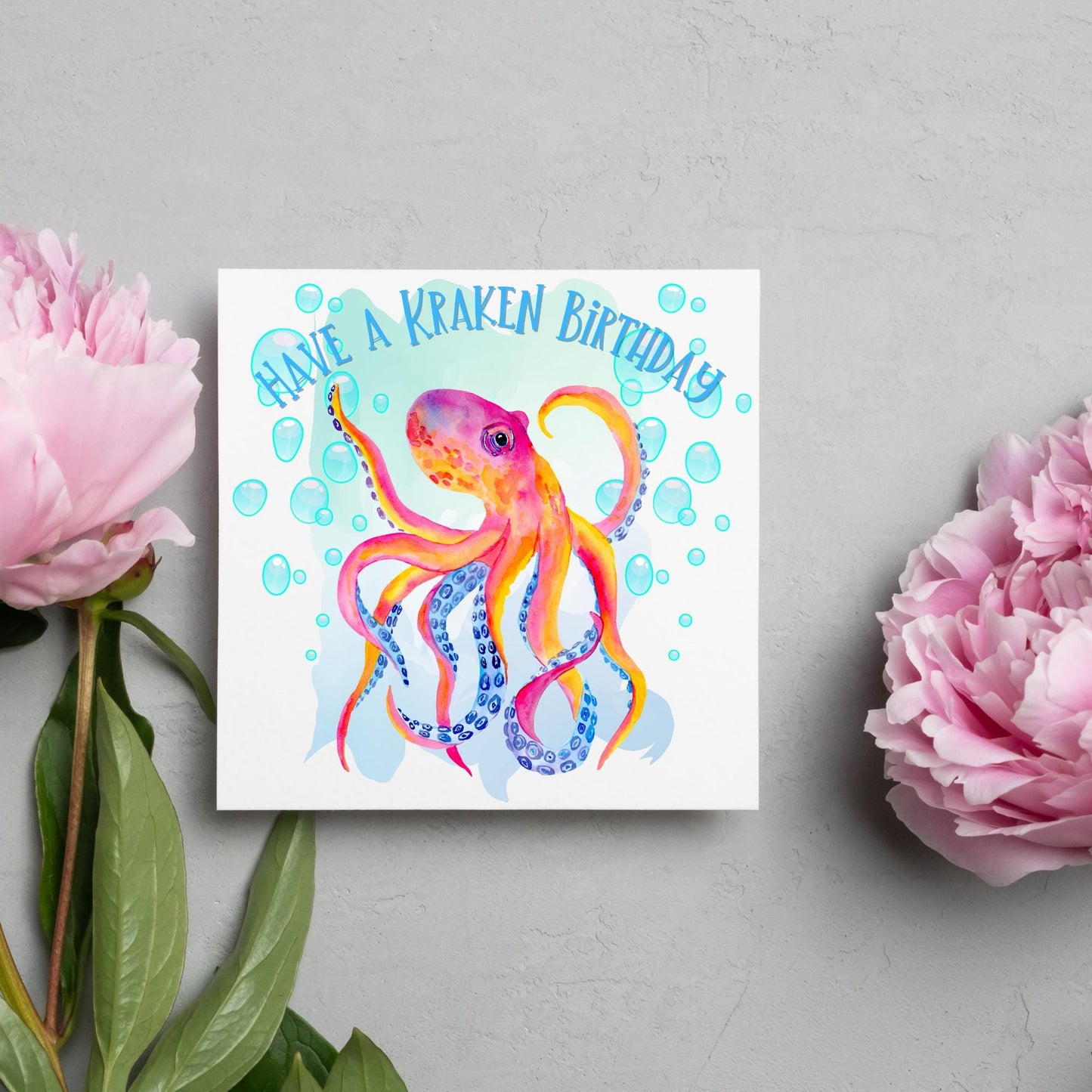 Image of a square birthday card. The background of the card is white and in the centre is a large watercolour image of an octopus in red, orange and blue tones against a light blue bubbly backdrop. Above the Octopus in blue capitalised text, have a kraken birthday is written in a handwritten style font
