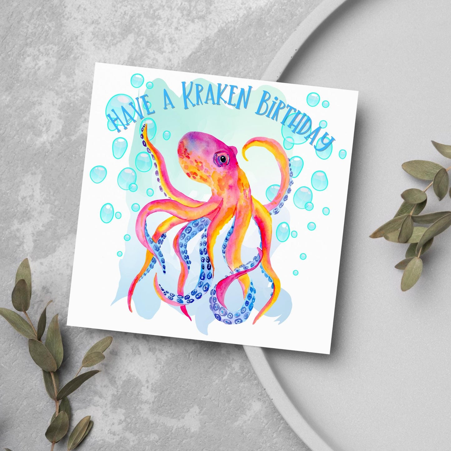 Image of a square birthday card. The background of the card is white and in the centre is a large watercolour image of an octopus in red, orange and blue tones against a light blue bubbly backdrop. Above the Octopus in blue capitalised text, have a kraken birthday is written in a handwritten style font