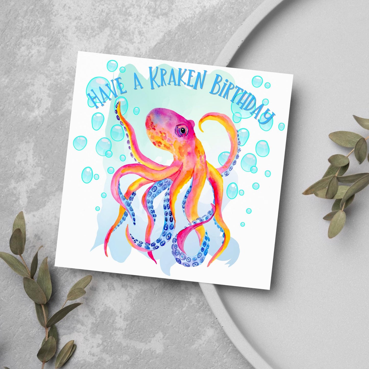 Image of a square birthday card. The background of the card is white and in the centre is a large watercolour image of an octopus in red, orange and blue tones against a light blue bubbly backdrop. Above the Octopus in blue capitalised text, have a kraken birthday is written in a handwritten style font