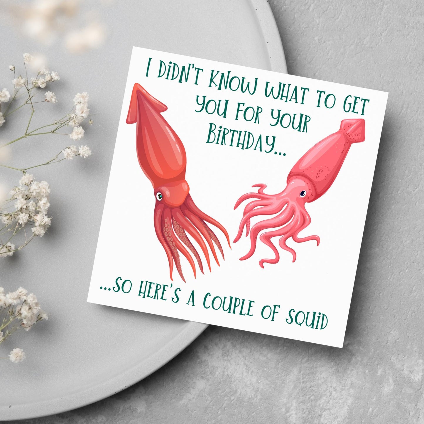 Image of a square birthday card. The background of the card is white and there is an illustration of 2 red squid in the centre. Writing surrounds the picture with 'I didn't know what to get you for your birthday....so here's a couple of squid' in a green capitalised handwritten font.
