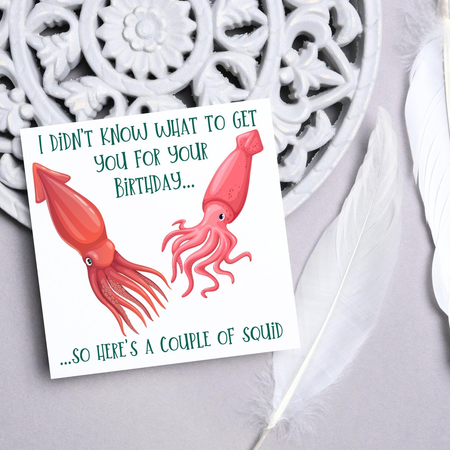 Image of a square birthday card. The background of the card is white and there is an illustration of 2 red squid in the centre. Writing surrounds the picture with 'I didn't know what to get you for your birthday....so here's a couple of squid' in a green capitalised handwritten font.