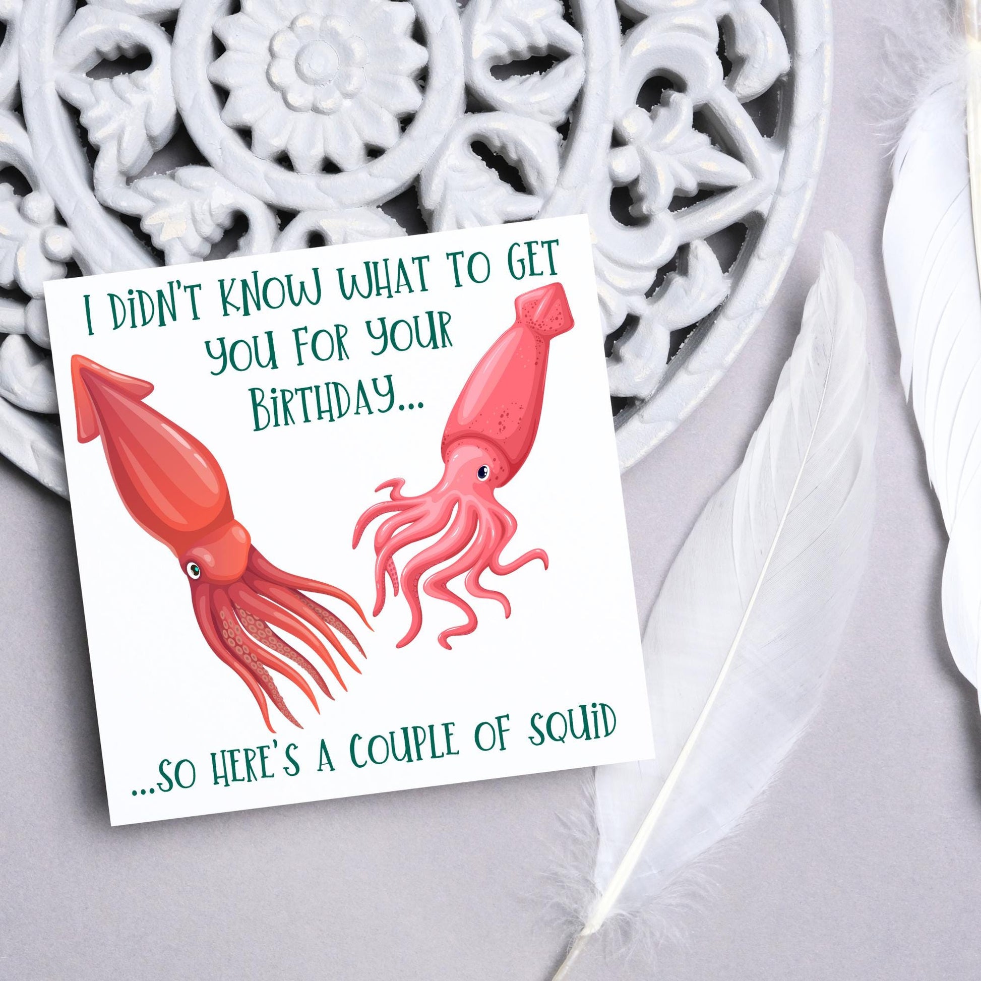 Image of a square birthday card. The background of the card is white and there is an illustration of 2 red squid in the centre. Writing surrounds the picture with 'I didn't know what to get you for your birthday....so here's a couple of squid' in a green capitalised handwritten font.