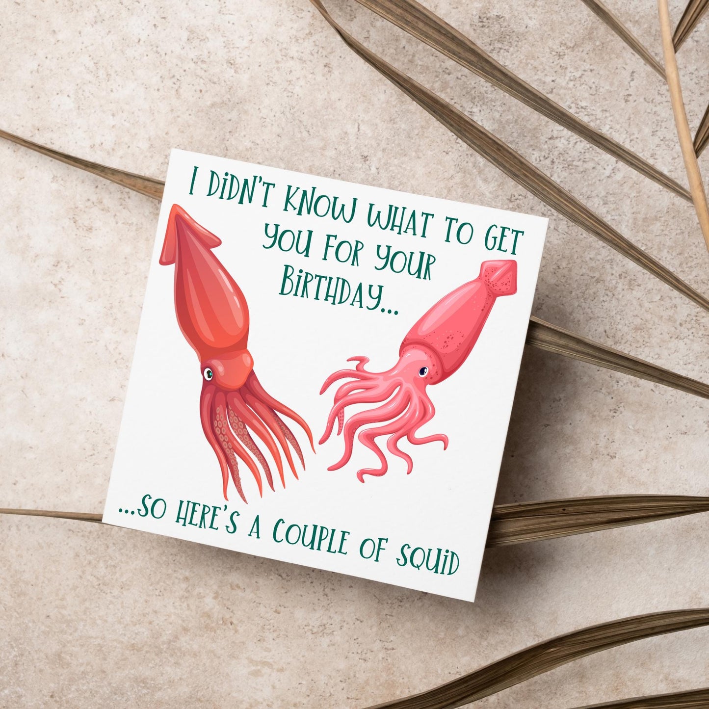 Image of a square birthday card. The background of the card is white and there is an illustration of 2 red squid in the centre. Writing surrounds the picture with 'I didn't know what to get you for your birthday....so here's a couple of squid' in a green capitalised handwritten font.