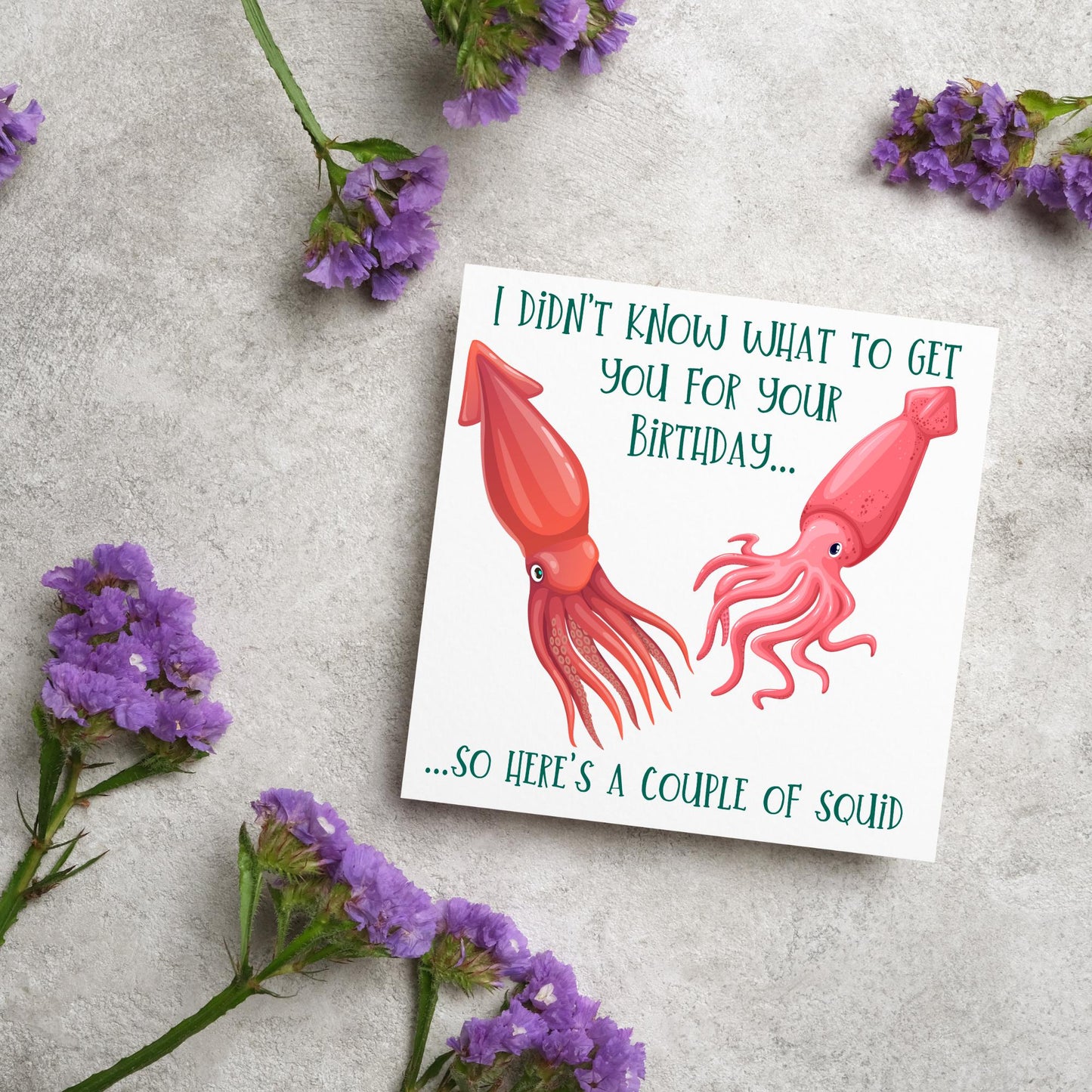 Image of a square birthday card. The background of the card is white and there is an illustration of 2 red squid in the centre. Writing surrounds the picture with 'I didn't know what to get you for your birthday....so here's a couple of squid' in a green capitalised handwritten font.