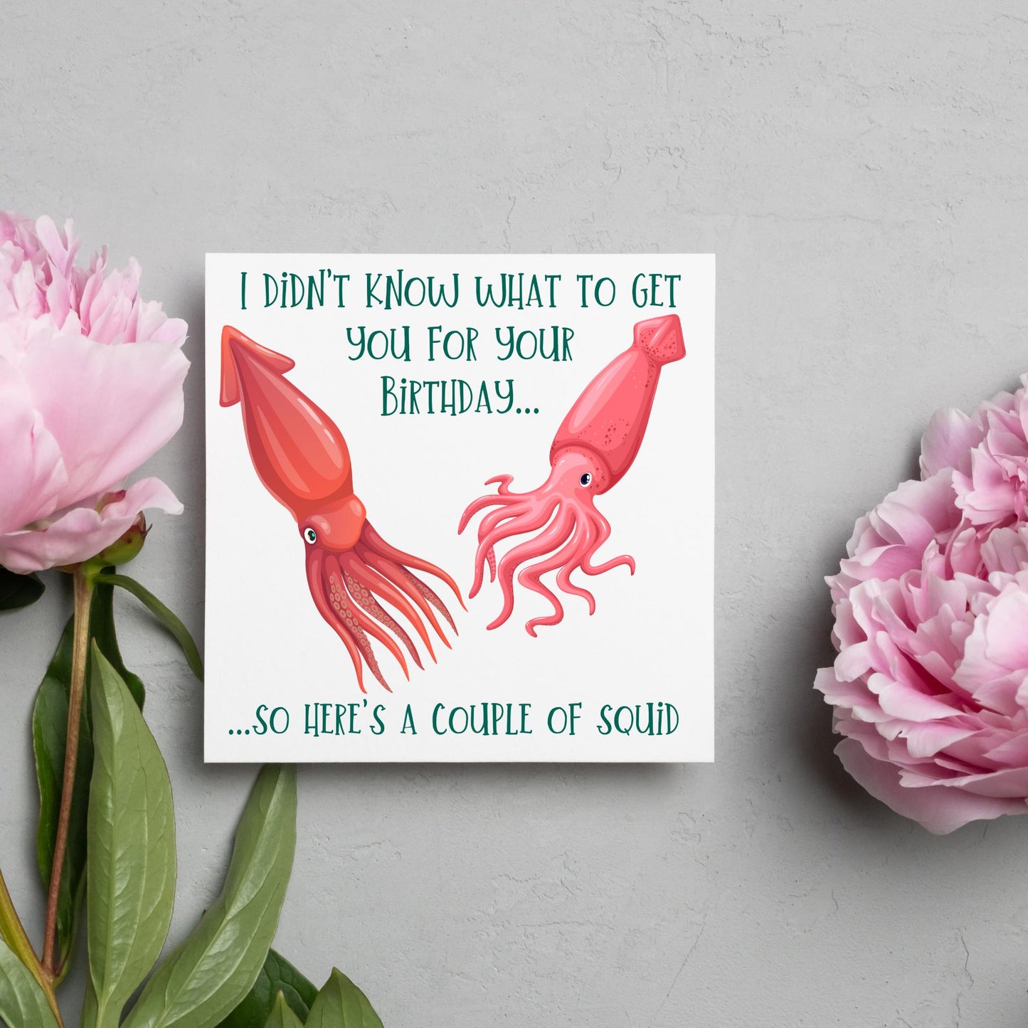 Image of a square birthday card. The background of the card is white and there is an illustration of 2 red squid in the centre. Writing surrounds the picture with 'I didn't know what to get you for your birthday....so here's a couple of squid' in a green capitalised handwritten font.