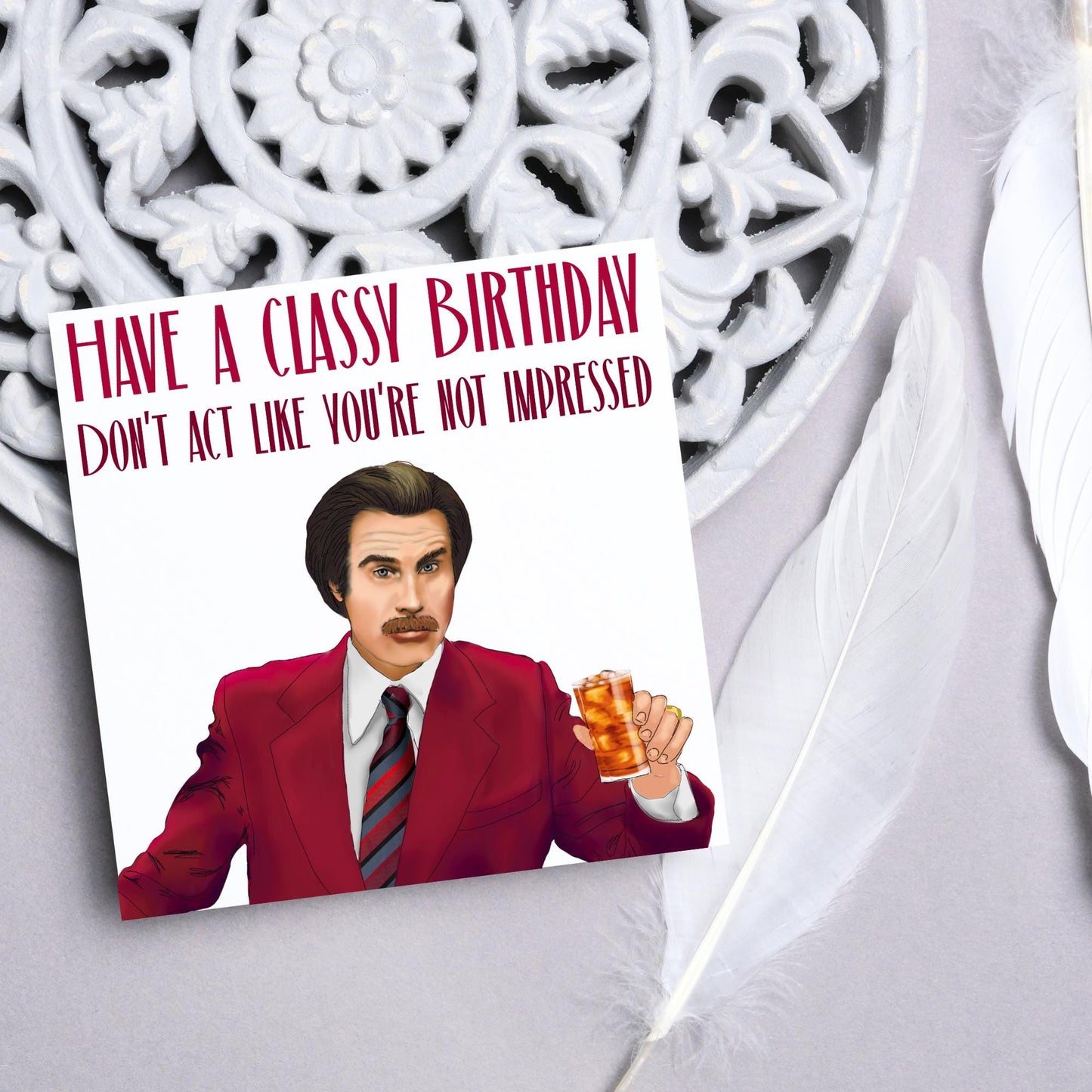 image of a square birthday card, the card has a white background with a hand drawn illustration of Ron Burgandy from the Movie Anchorman in a dark red suit holding a glass of whiskey. Above him is dark red capitalised text that reads 'Have a classy birthday, Don't act like you're not impressed.'