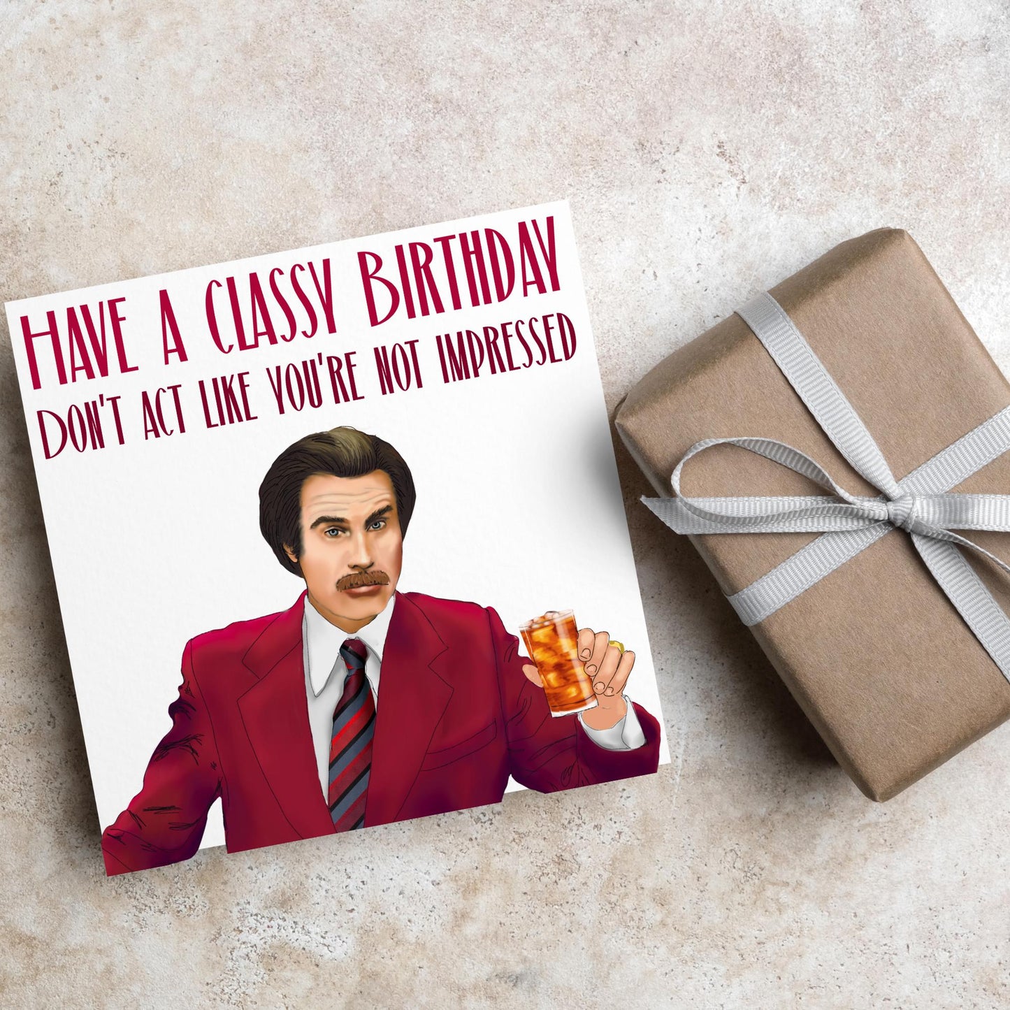 image of a square birthday card, the card has a white background with a hand drawn illustration of Ron Burgandy from the Movie Anchorman in a dark red suit holding a glass of whiskey. Above him is dark red capitalised text that reads 'Have a classy birthday, Don't act like you're not impressed.'