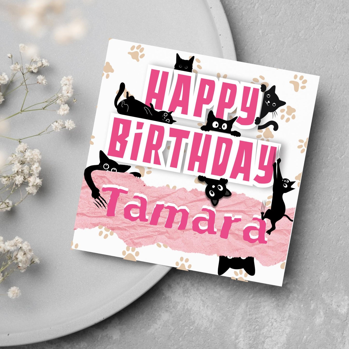 A square birthday card with a white background featuring playful black cartoon cats surrounding and interacting with bold pink text that says "HAPPY BiRTHDAY." The name "Tamara" is written in a casual, pink handwritten font across a torn paper-style pink banner. Light brown paw prints are scattered in the background.