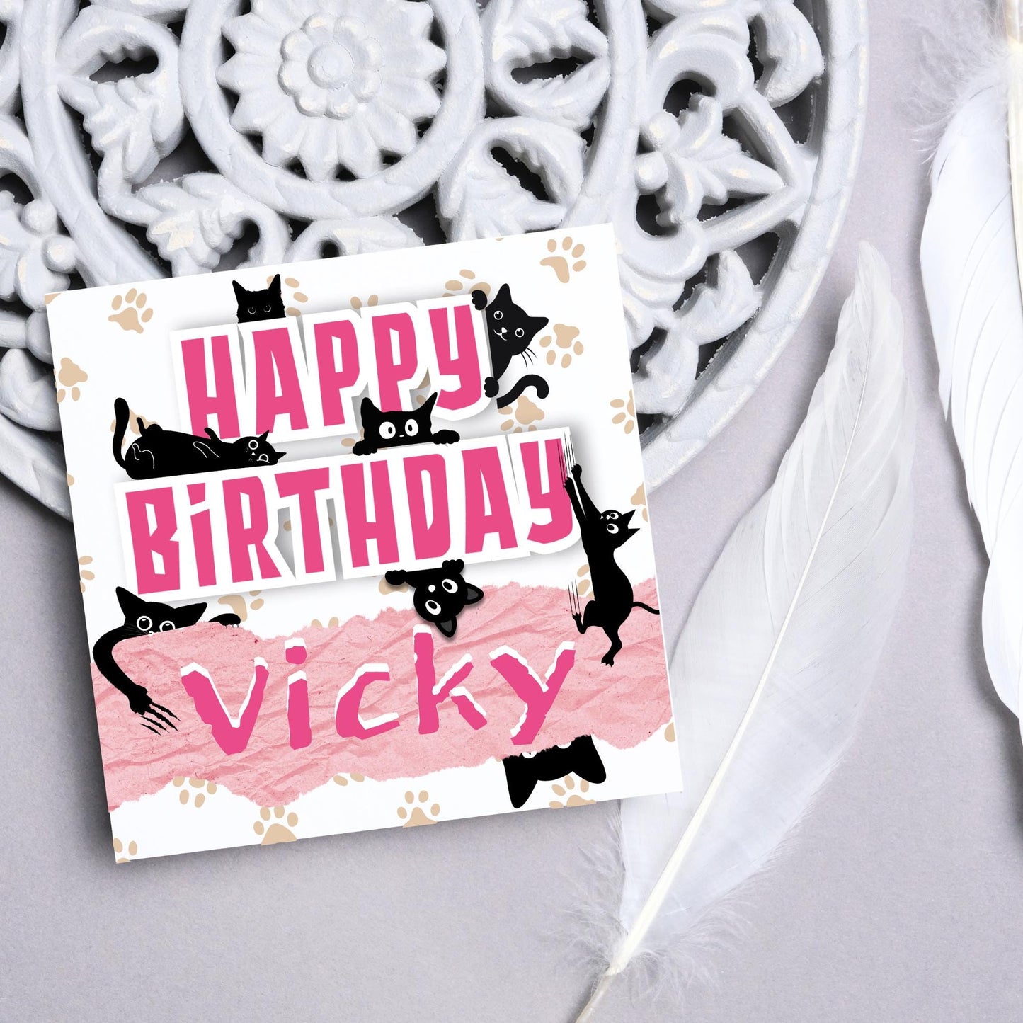 A square birthday card with a white background featuring playful black cartoon cats surrounding and interacting with bold pink text that says "HAPPY BiRTHDAY." The name "Vicky" is written in a casual, pink handwritten font across a torn paper-style pink banner. Light brown paw prints are scattered in the background.