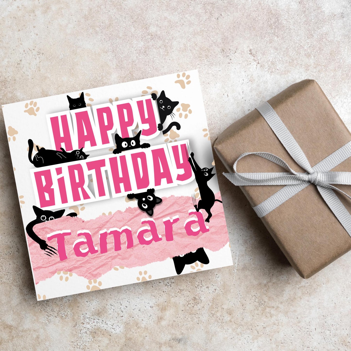 A square birthday card with a white background featuring playful black cartoon cats surrounding and interacting with bold pink text that says "HAPPY BiRTHDAY." The name "Tamara" is written in a casual, pink handwritten font across a torn paper-style pink banner. Light brown paw prints are scattered in the background.