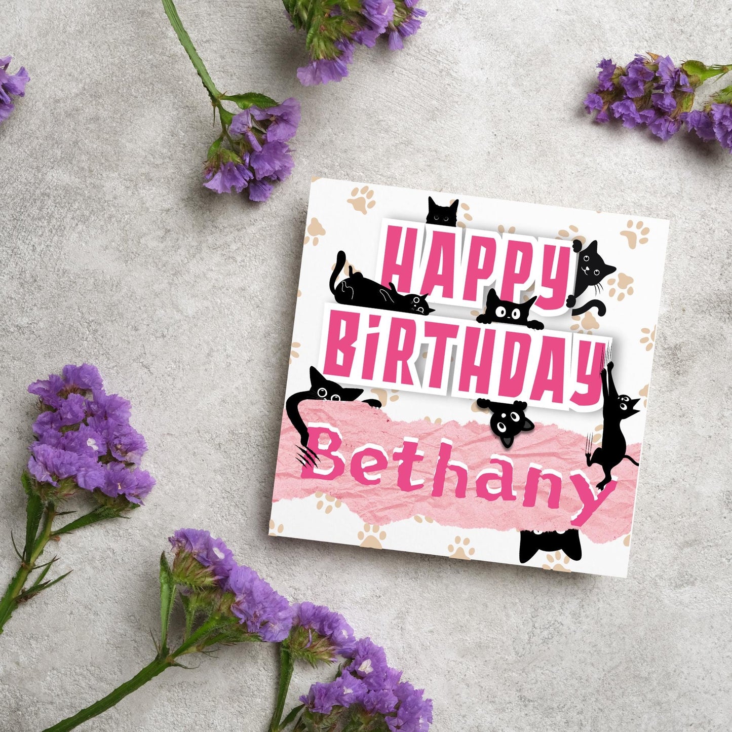 A square birthday card with a white background featuring playful black cartoon cats surrounding and interacting with bold pink text that says "HAPPY BiRTHDAY." The name "Bethany" is written in a casual, pink handwritten font across a torn paper-style pink banner. Light brown paw prints are scattered in the background.