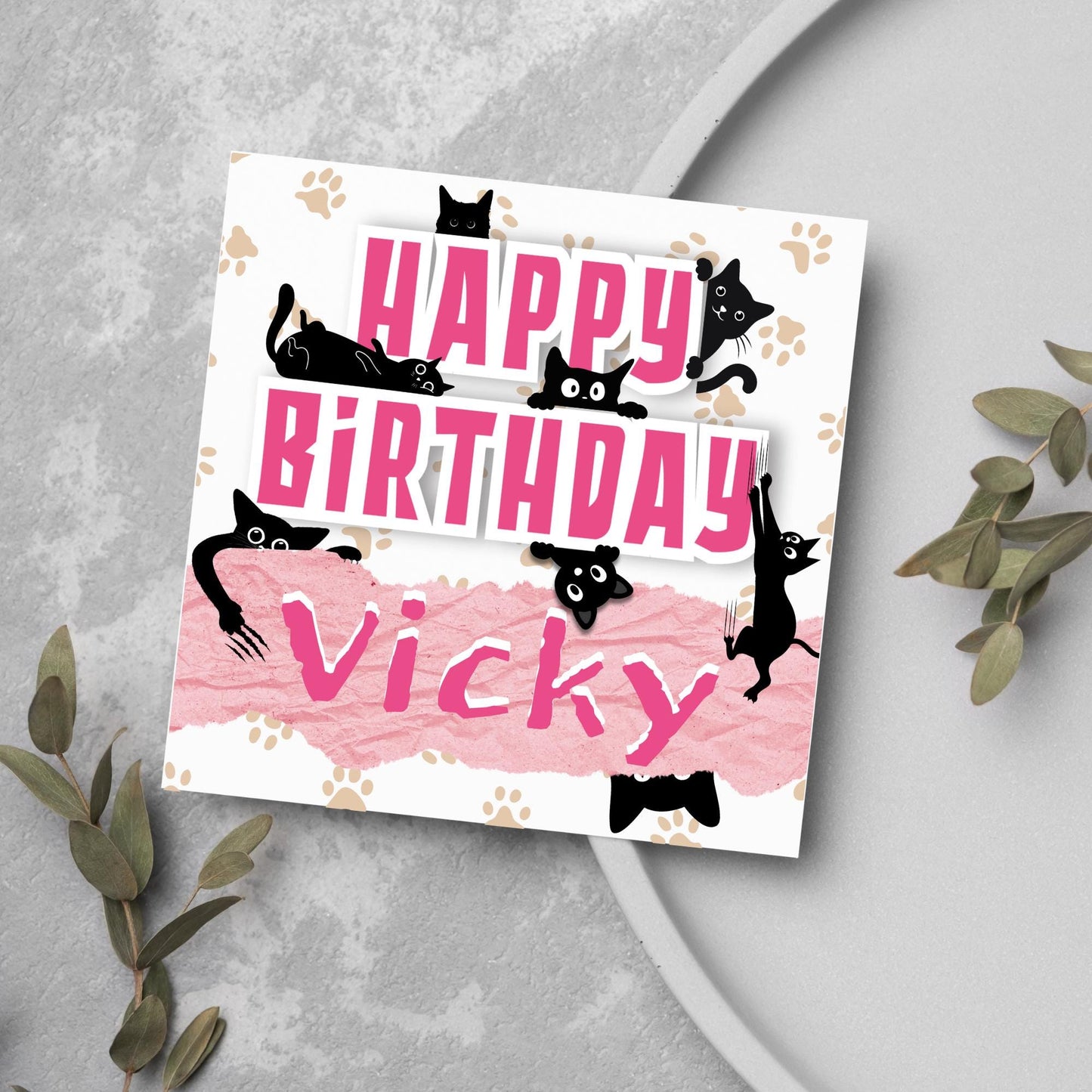 A square birthday card with a white background featuring playful black cartoon cats surrounding and interacting with bold pink text that says "HAPPY BiRTHDAY." The name "Vicky" is written in a casual, pink handwritten font across a torn paper-style pink banner. Light brown paw prints are scattered in the background.