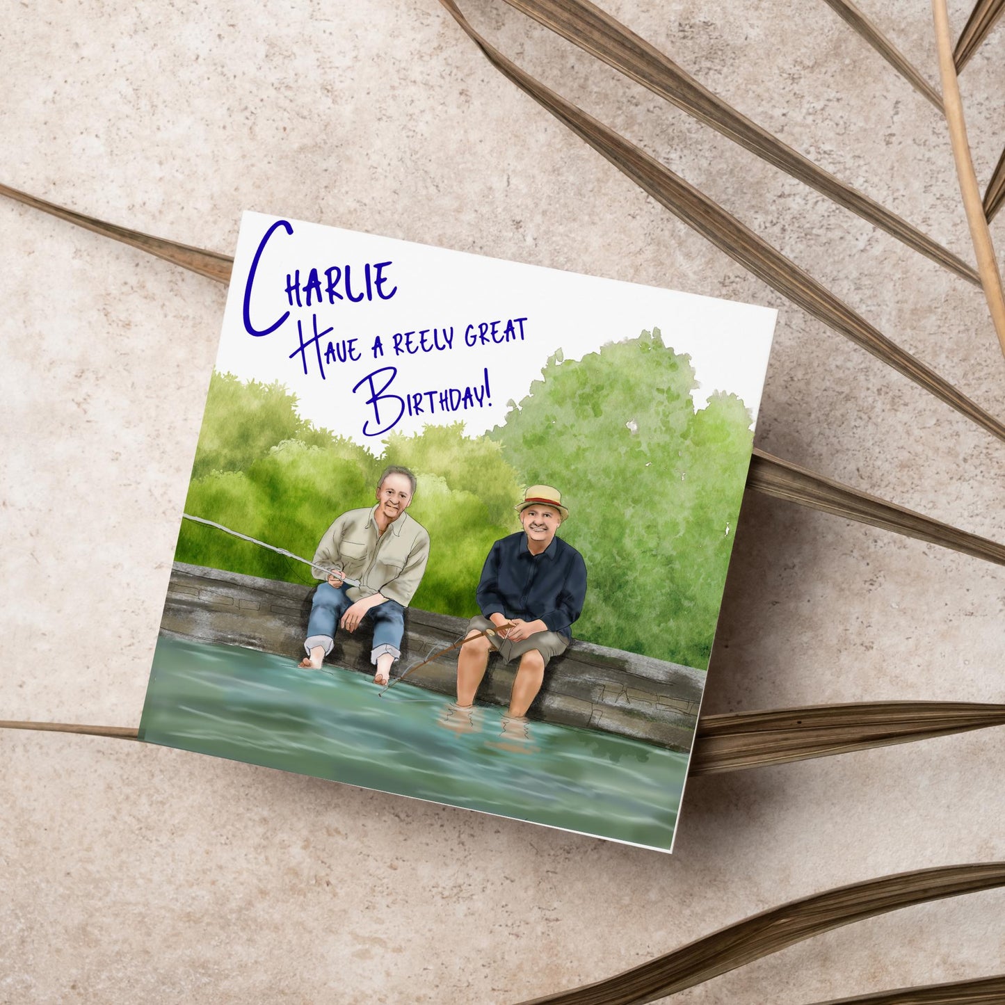 square greetings card with a hand drawn image of bob Mortimer and paul Whitehouse sitting on the edge of a dock fishing with their bare feet in the water. behind them are watercolour trees and bushes and blue text that reads 'charlie, have a freely great birthday' in a handwritten style font