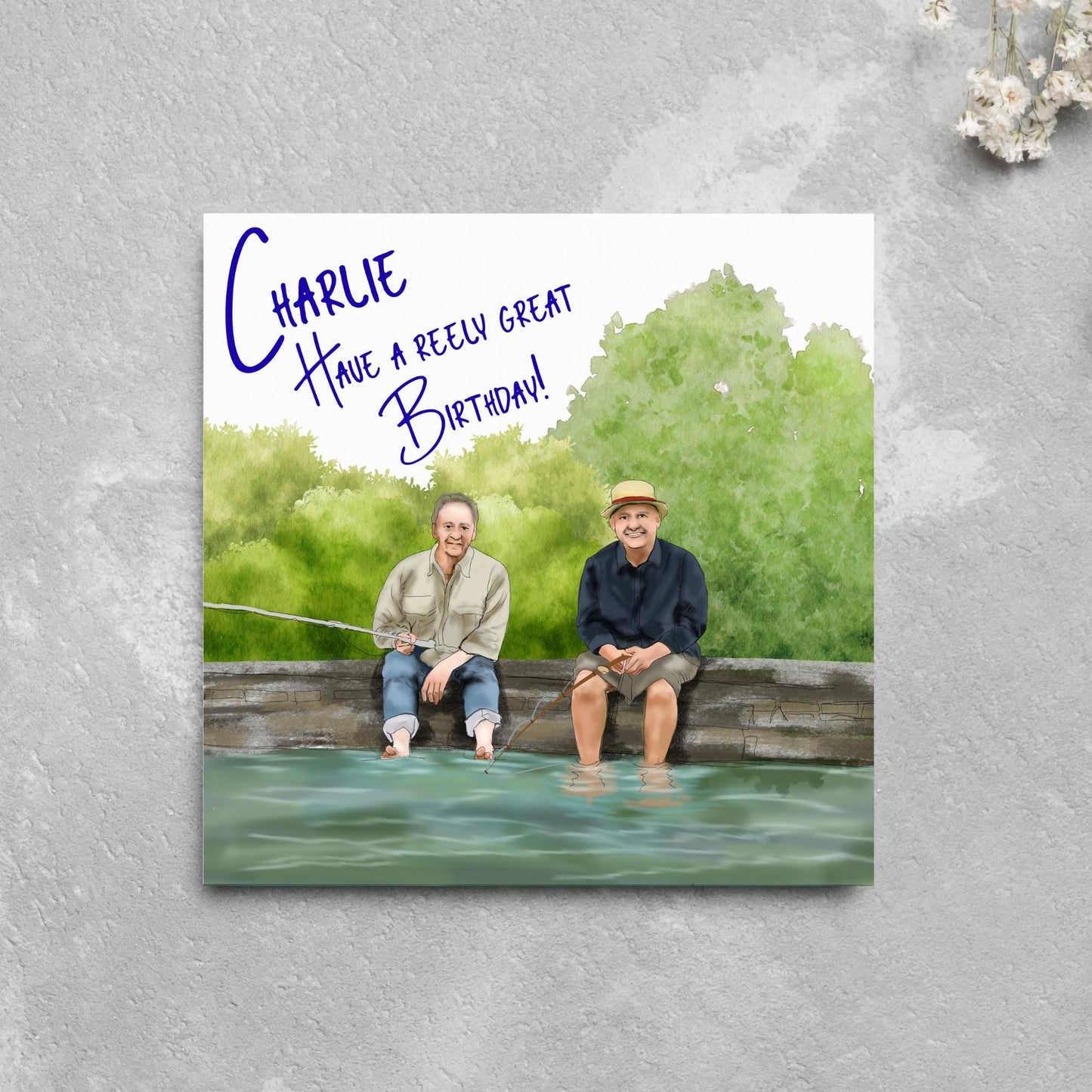 square greetings card with a hand drawn image of bob Mortimer and paul Whitehouse sitting on the edge of a dock fishing with their bare feet in the water. behind them are watercolour trees and bushes and blue text that reads 'charlie, have a freely great birthday' in a handwritten style font