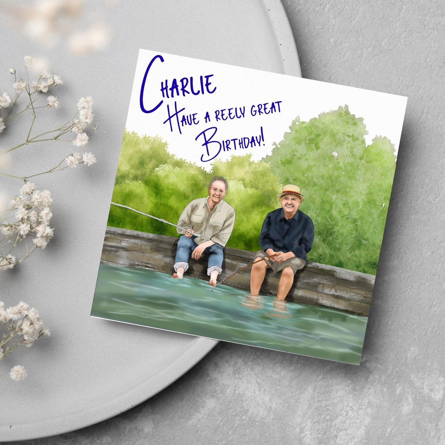 square greetings card with a hand drawn image of bob Mortimer and paul Whitehouse sitting on the edge of a dock fishing with their bare feet in the water. behind them are watercolour trees and bushes and blue text that reads 'charlie, have a freely great birthday' in a handwritten style font