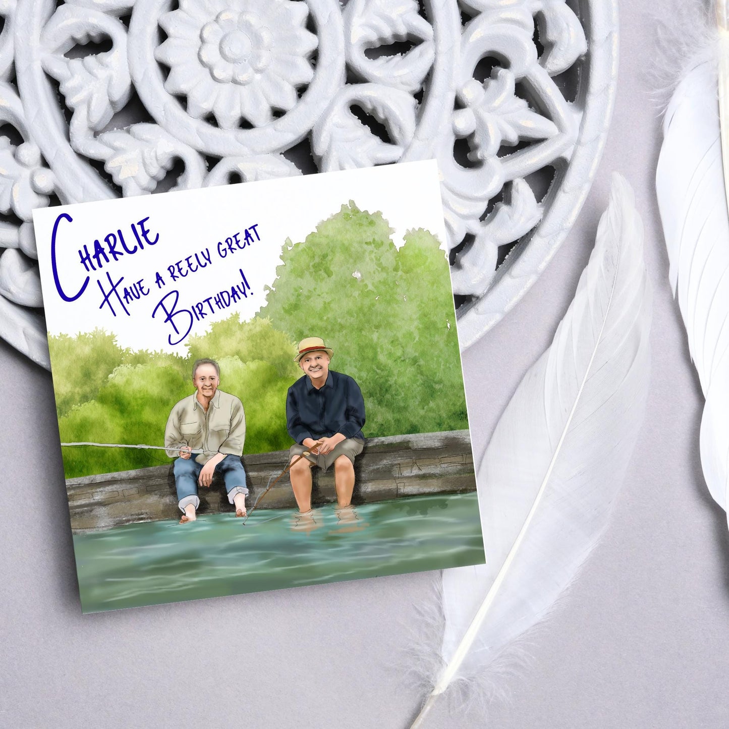 square greetings card with a hand drawn image of bob Mortimer and paul Whitehouse sitting on the edge of a dock fishing with their bare feet in the water. behind them are watercolour trees and bushes and blue text that reads 'charlie, have a freely great birthday' in a handwritten style font