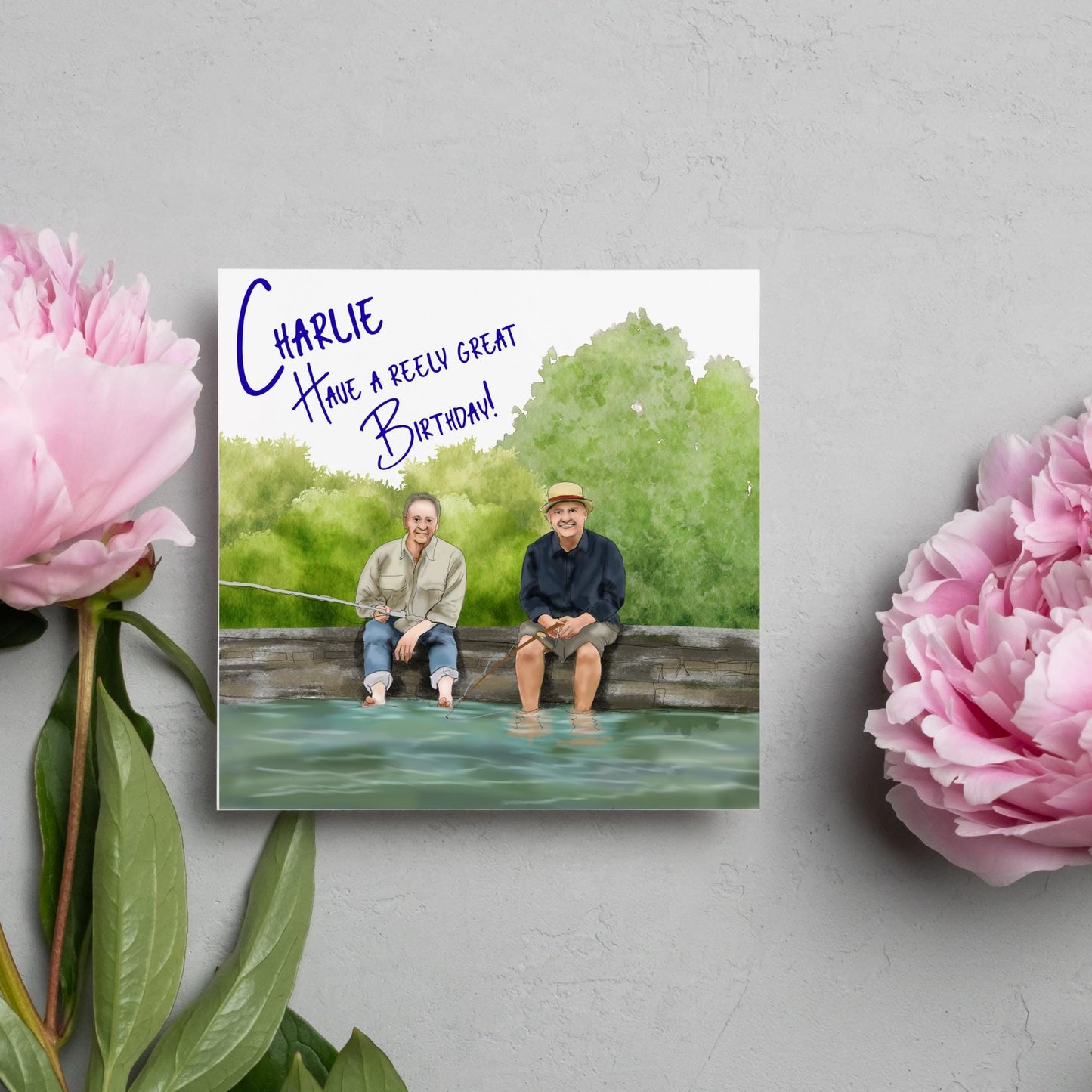 square greetings card with a hand drawn image of bob Mortimer and paul Whitehouse sitting on the edge of a dock fishing with their bare feet in the water. behind them are watercolour trees and bushes and blue text that reads 'charlie, have a freely great birthday' in a handwritten style font