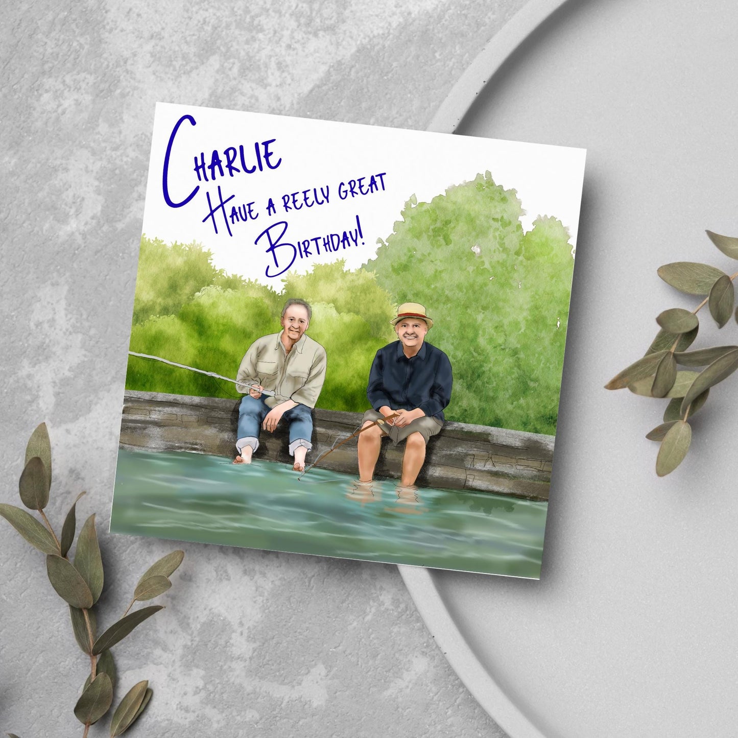square greetings card with a hand drawn image of bob Mortimer and paul Whitehouse sitting on the edge of a dock fishing with their bare feet in the water. behind them are watercolour trees and bushes and blue text that reads 'charlie, have a freely great birthday' in a handwritten style font