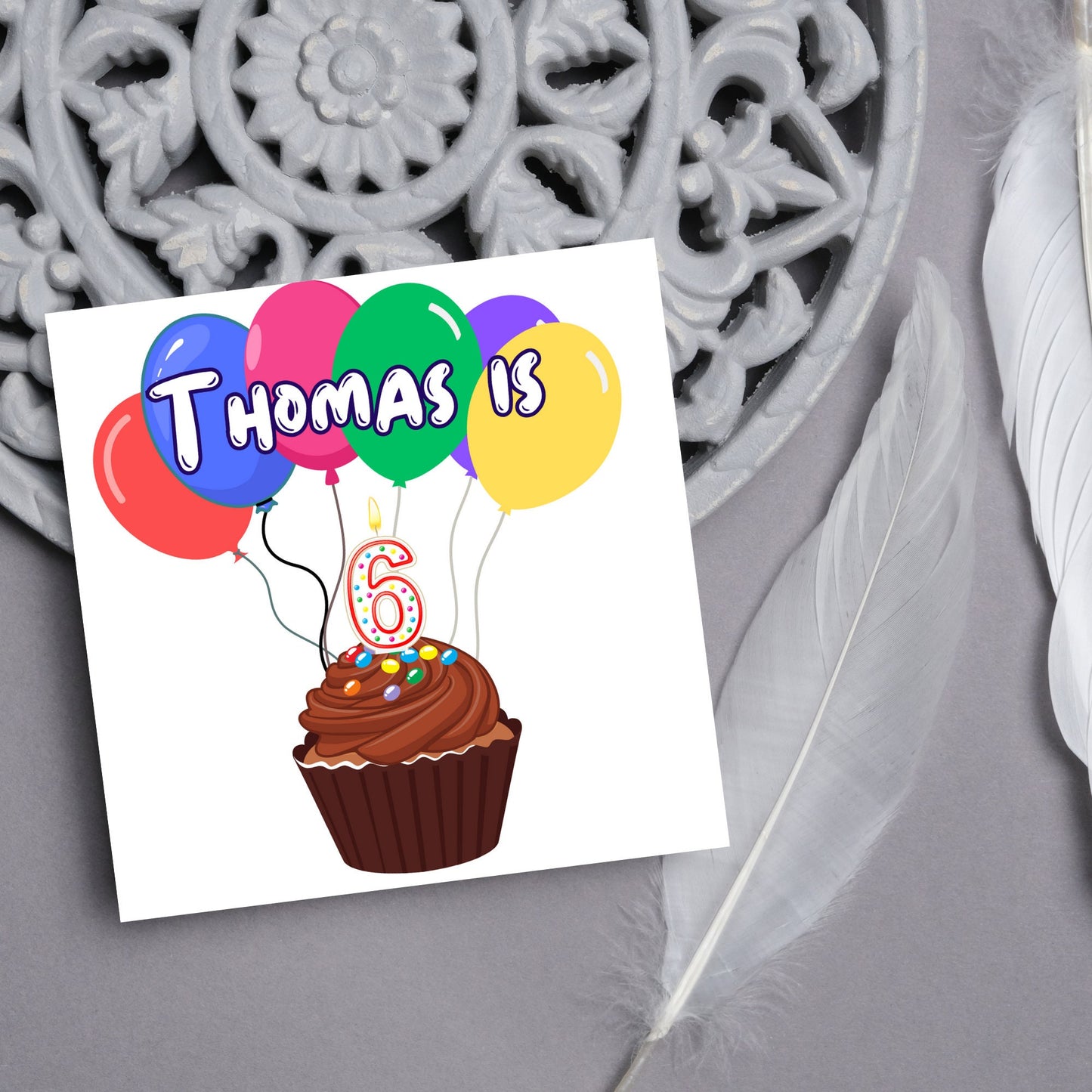 square childs birthday card with chocolate cupcake design personalised with name balloons and a number 6 candle. card reads thomas is 6
