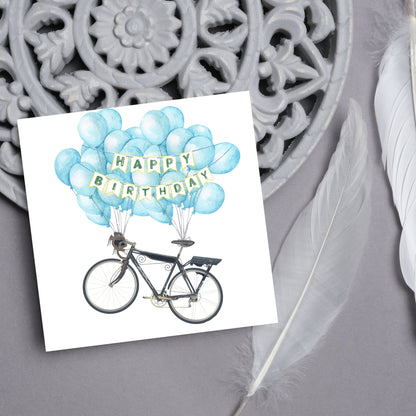 White square birthday card with an illustration of a bicycle being held aloft by blue watercolour balloons and bunting that reads happy birthday