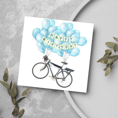 White square birthday card with an illustration of a bicycle being held aloft by blue watercolour balloons and bunting that reads happy birthday