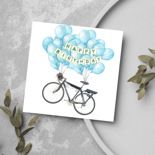 White square birthday card with an illustration of a bicycle being held aloft by blue watercolour balloons and bunting that reads happy birthday