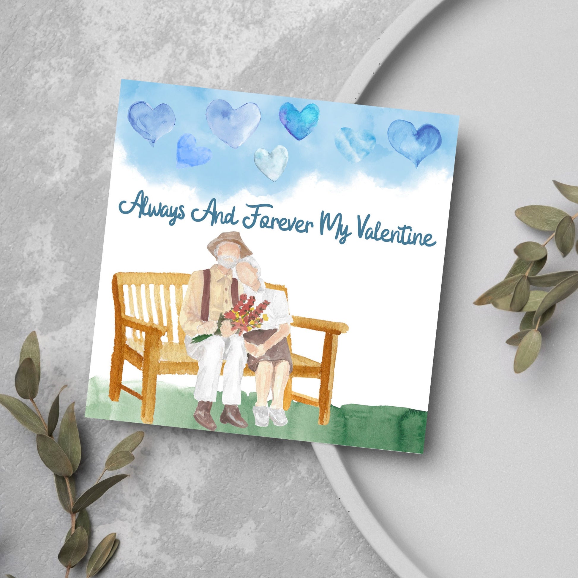 This beautiful Card has a watercolour style print with a couple sitting on a bench with flowers. Above them the blue sky has a series of cute watercolour hearts. This Cute card is perfect for valentines day for your wife or husband